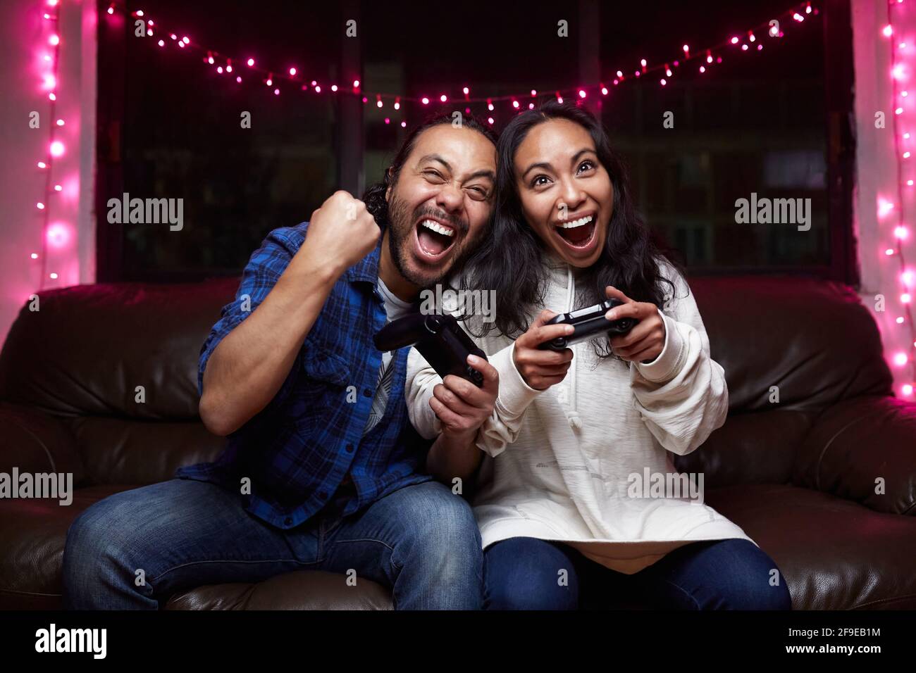 Aufgeregtes ethnisches Paar in Freizeitkleidung mit spielenden Joy Pads Videospiel zusammen, während Sie zu Hause auf der Ledercouch sitzen Stockfoto