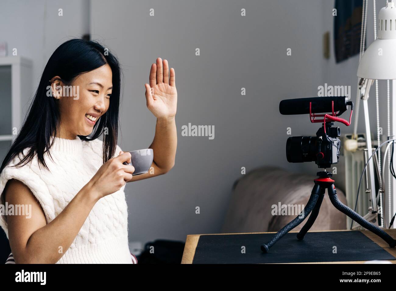 Lächelnde ethnische Bloggerin mit einer Tasse Heißgetränk hallo Geste während der Aufnahme von Video auf Fotokamera zu Hause Stockfoto