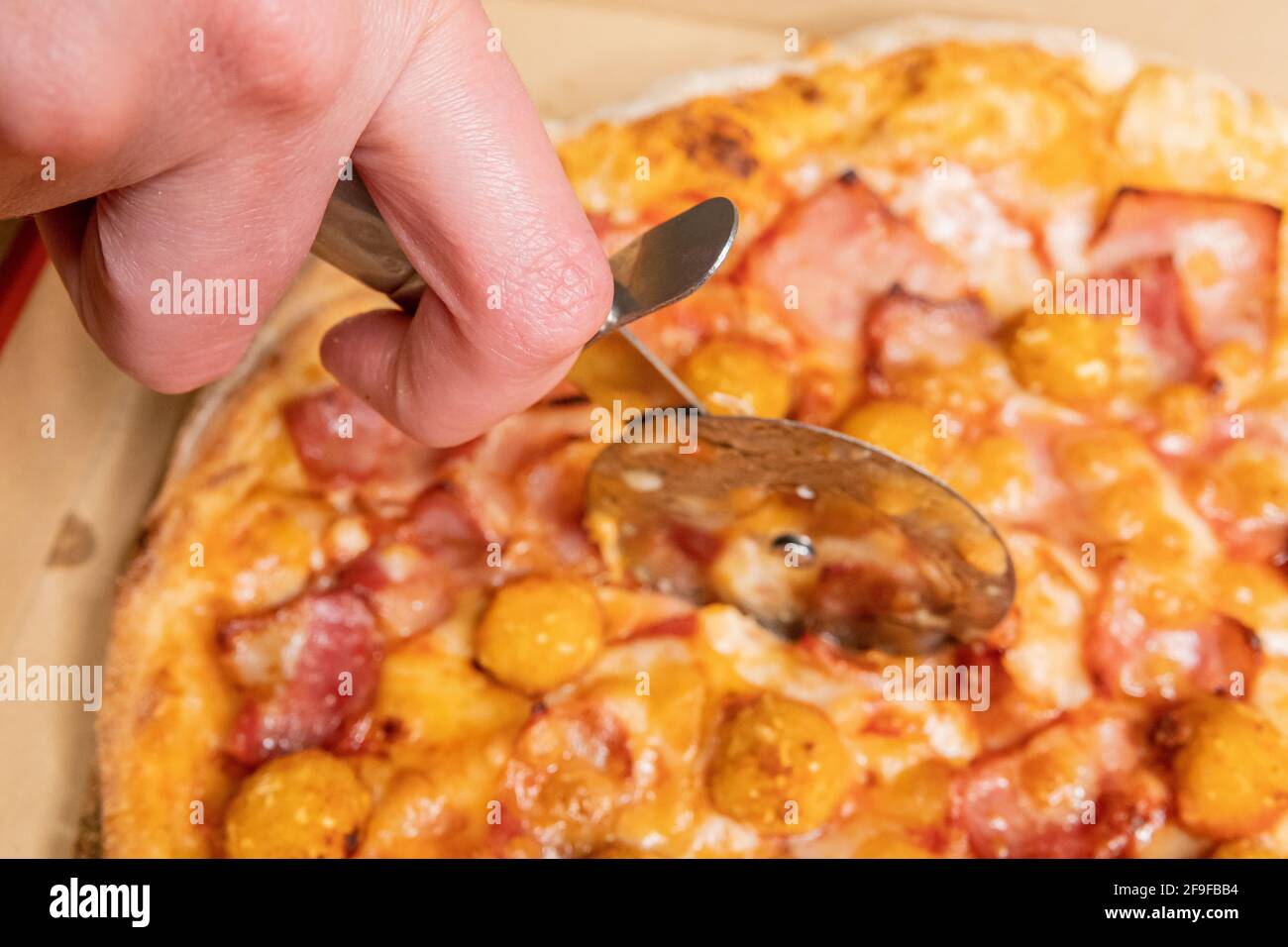 Nahaufnahme einer Hand mit einem Pizzaschneider Eine Pizza mit Speck und Huhn knallt auf eine Schachtel Stockfoto