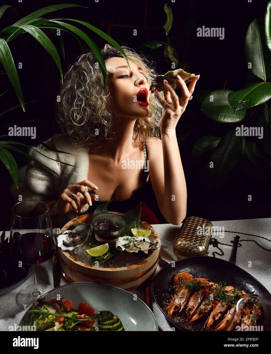 Die junge blonde Frau im Abendkleid mit tiefem Ausschnitt isst im Restaurant Meeresfrüchte-Austern, Garnelen, Muscheln und Salat Stockfoto