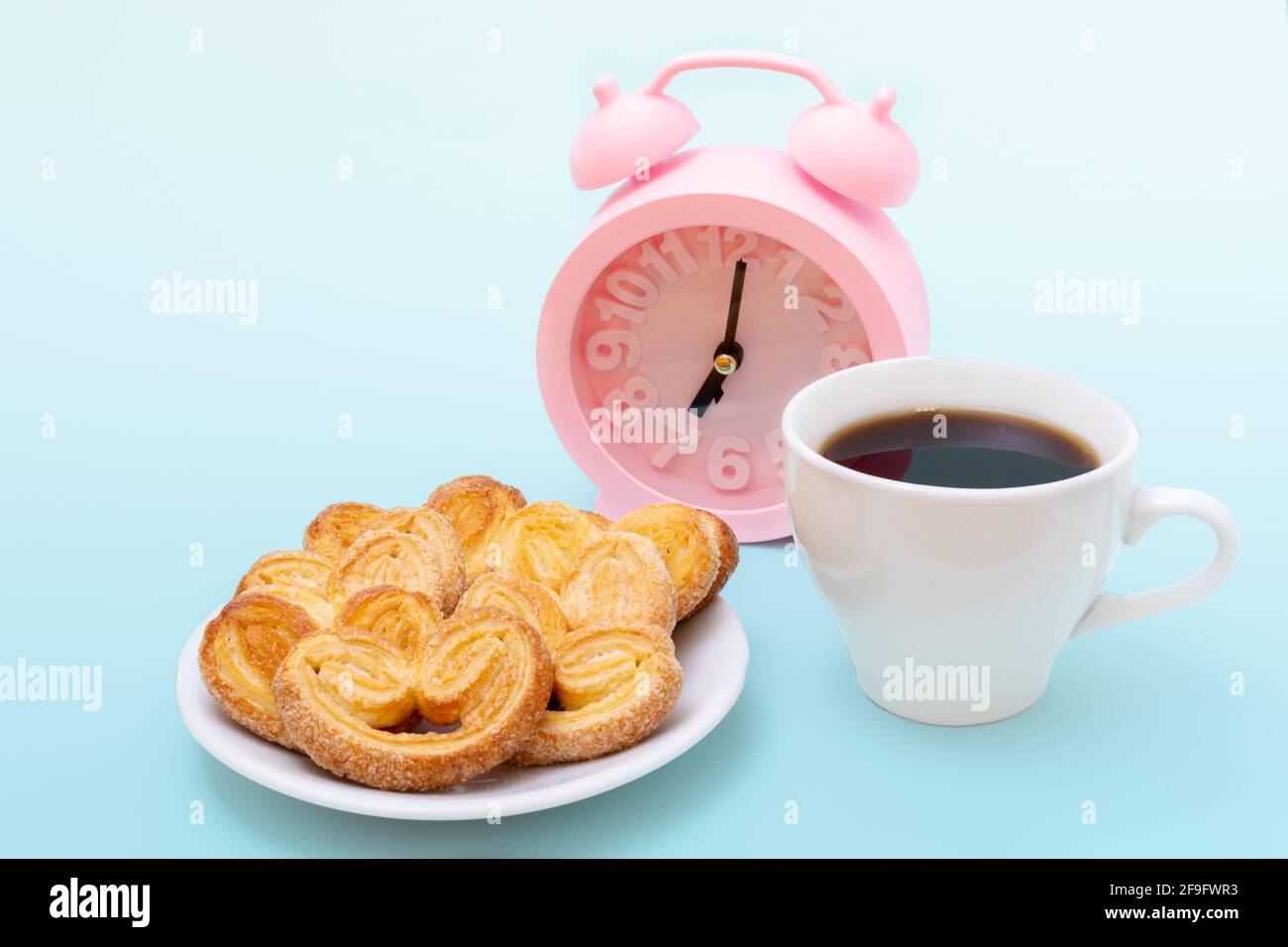 Weiße Tasse dampfenden heißen schwarzen Kaffee oder heiße Schokolade, frisch gebackene herzförmige Kekse und rosa Wecker auf hellblauem Hintergrund, Kopierraum. Stockfoto
