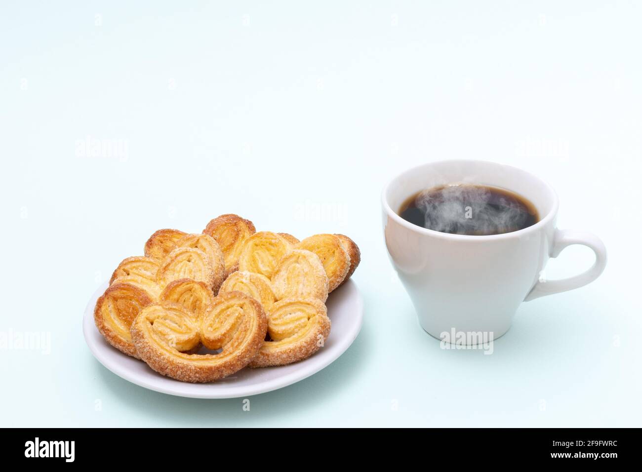 Weiße Tasse dampfenden heißen schwarzen Kaffee oder heiße Schokolade und frisch gekochte herzförmige Kekse auf hellblauem Hintergrund, Platz zum Kopieren. Frühstück am Morgen Stockfoto