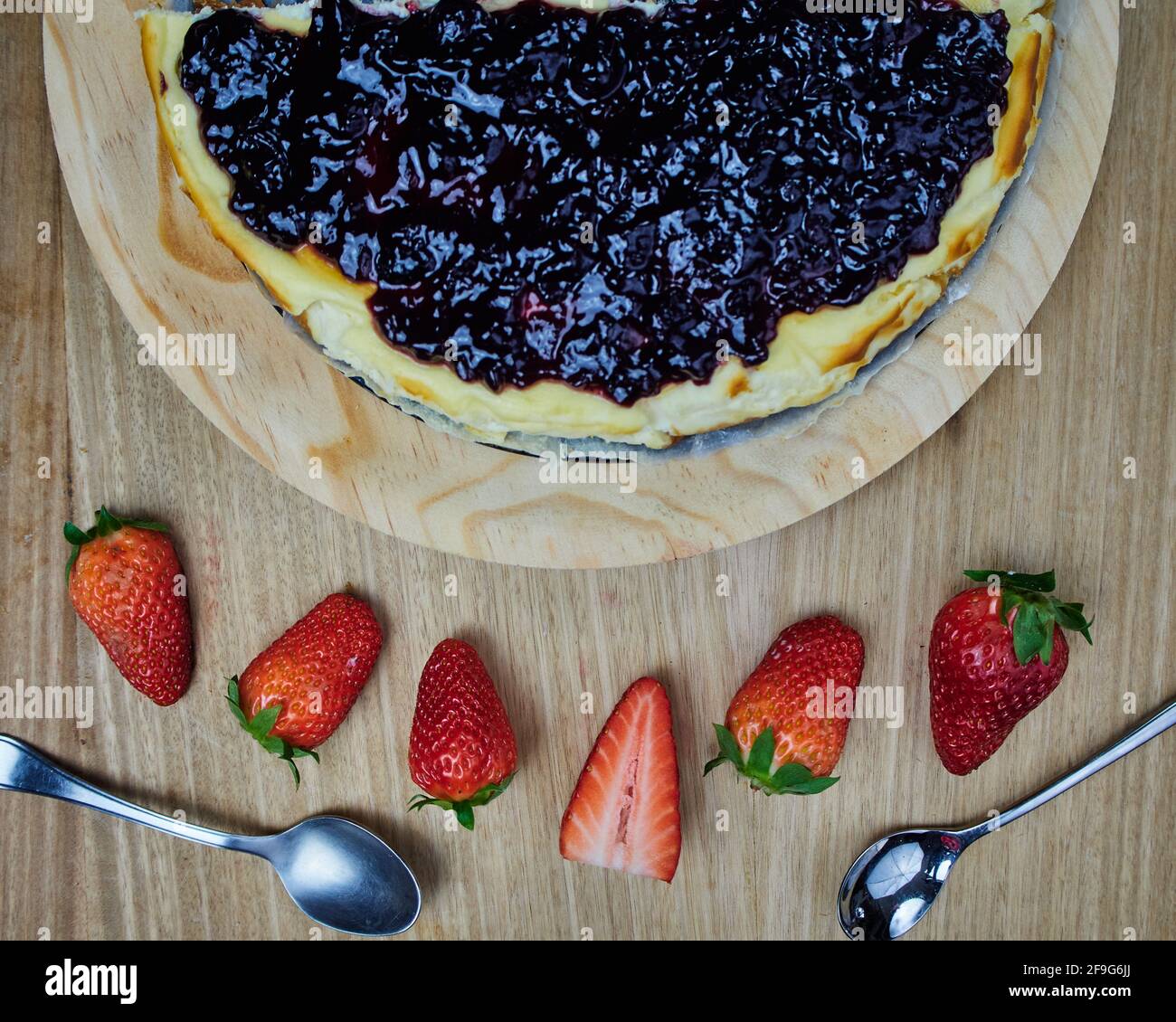 Die Hälfte des Käsekuchen auf einem runden Holzteller und mehrere Erdbeeren und zwei Löffel. Draufsicht Stockfoto