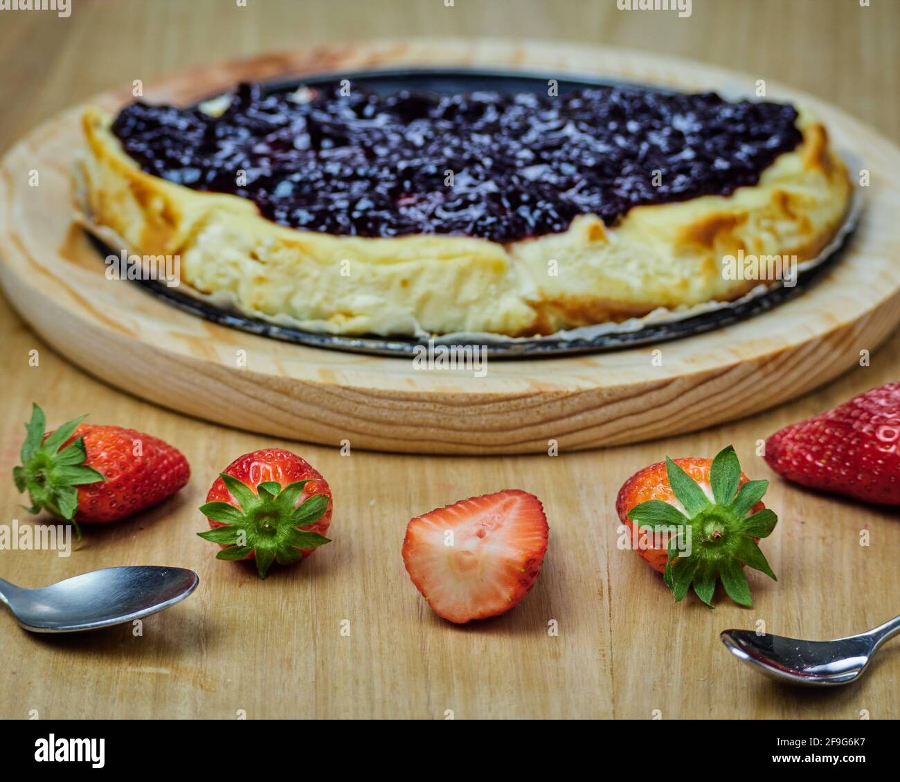 Die Hälfte des Käsekuchen auf einem runden Holzteller und mehrere Erdbeeren und zwei Löffel. Stockfoto