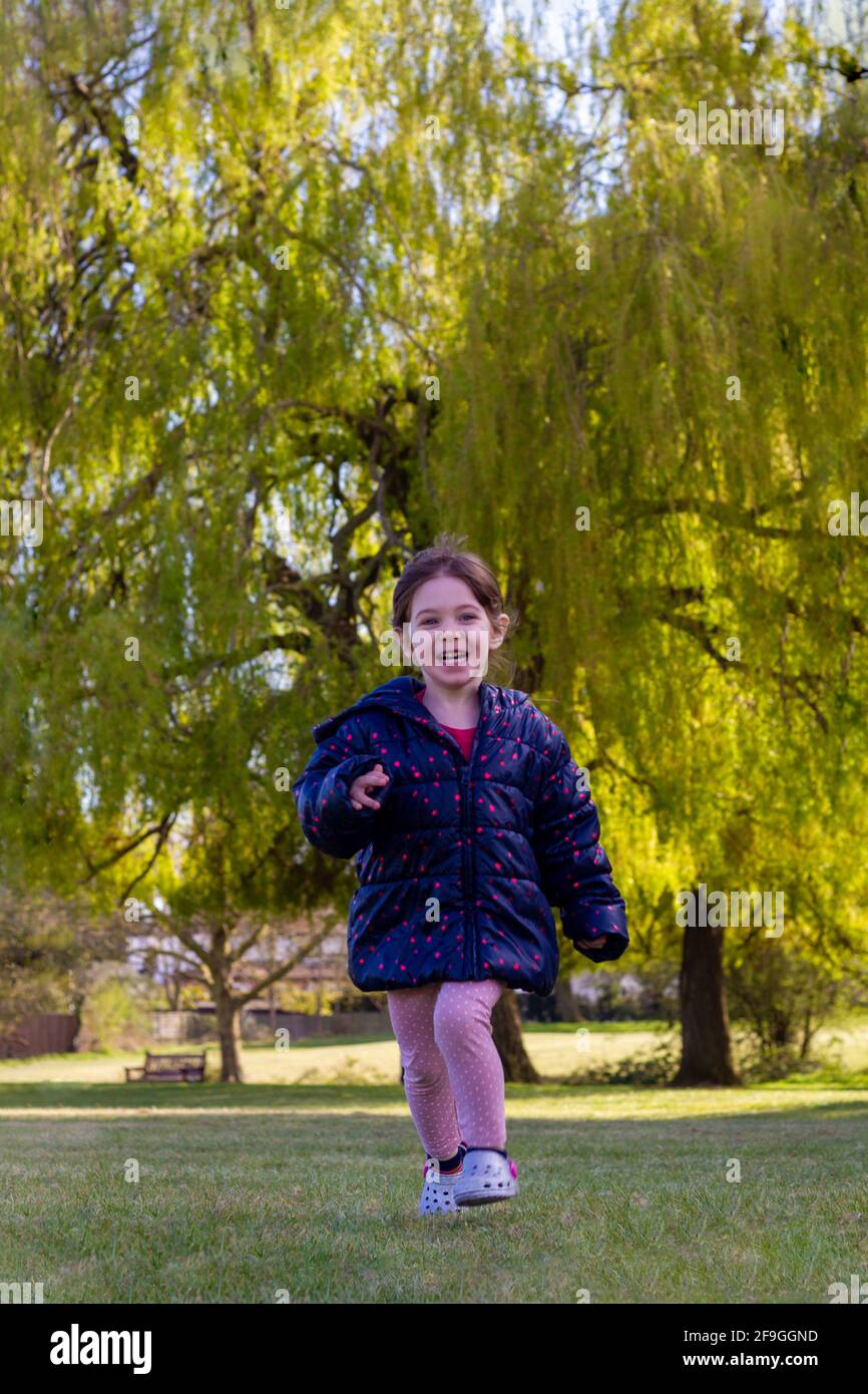 Ganzkörperaufnahme eines niedlichen braunen Haarbabys mit Eine blaue Jacke und eine rosa Hose, die auf dem Gras läuft In einem Park an einem sonnigen Frühlingstag Stockfoto