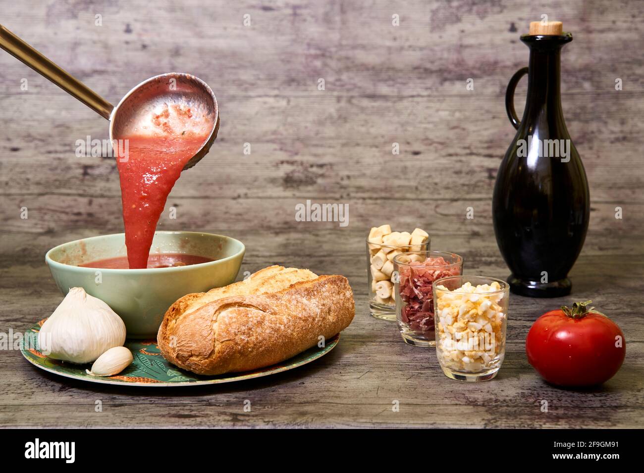 Gazpacho, der in die Schüssel fällt und serviert wird. Das Gericht wird von Brot und Knoblauch begleitet, und auf der rechten Seite sind Croutons, Ei und Serrano-Schinken, ein tomat Stockfoto