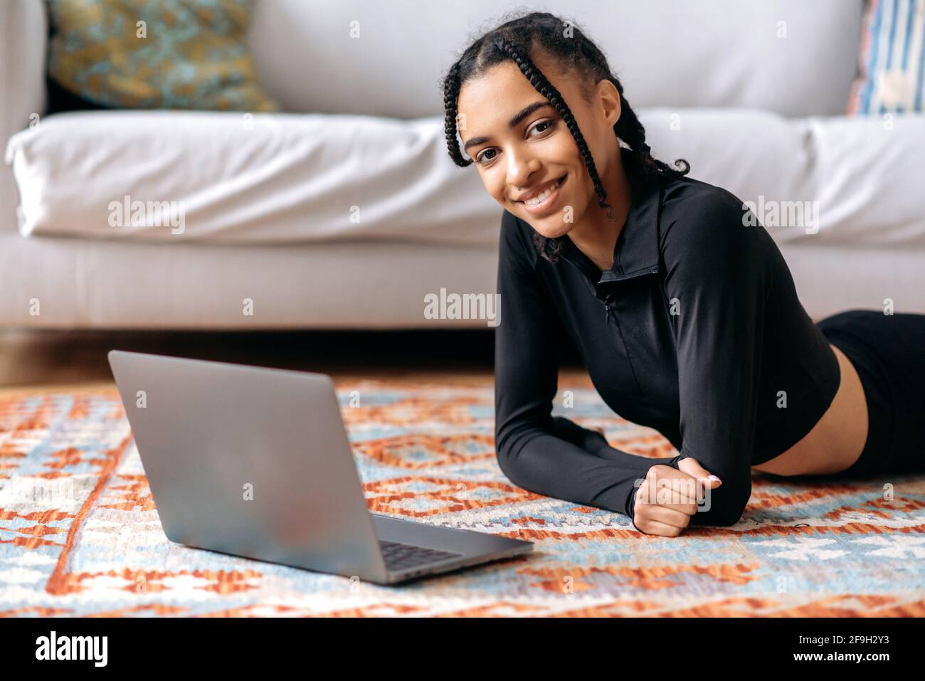 Sportlich schöne junge afroamerikanische Frau mit einer guten Figur, in Sportkleidung, liegt zu Hause auf dem Boden, Uhren Online-Fitnessunterricht nutzt Laptop, geht in den Sport, führt einen gesunden Lebensstil Stockfoto