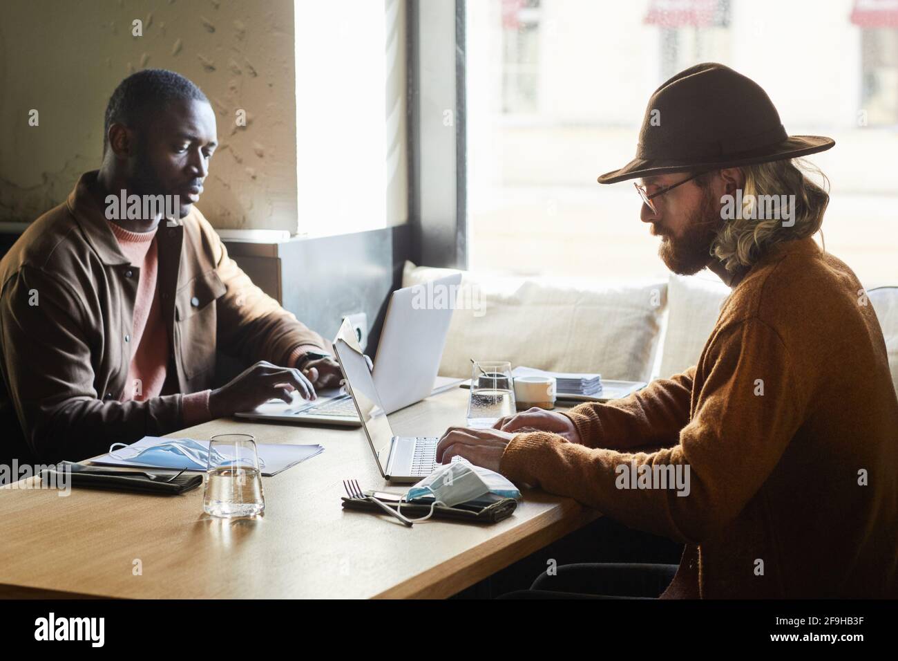 Porträt von zwei zeitgenössischen Männern, die Laptops benutzen, während sie an einem von Sonnenlicht beleuchteten Kaffeetisch im Loft arbeiten, Kopierraum Stockfoto