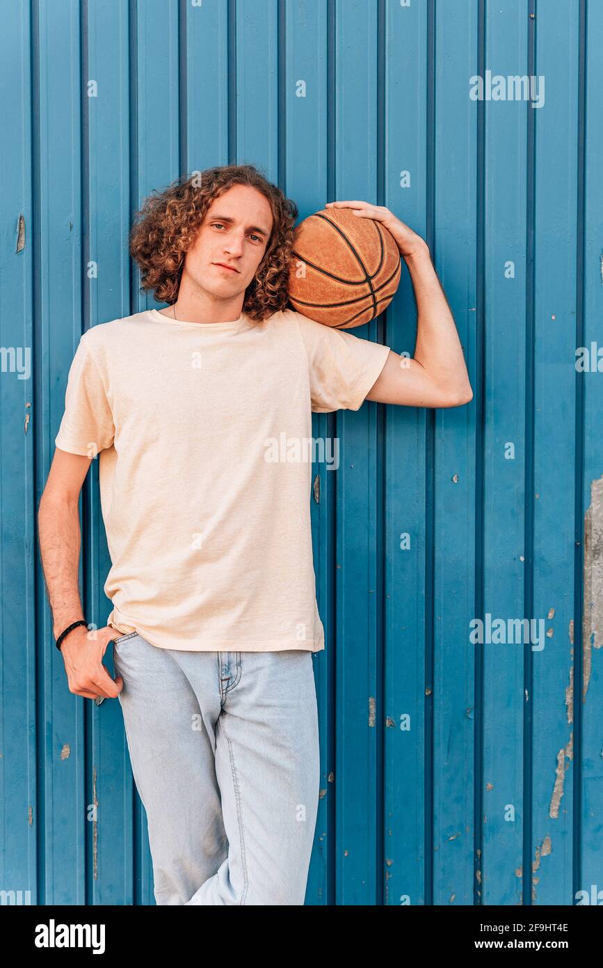 Vertikales Porträt eines jungen kaukasischen Mannes, der einen Basketball auf der Schulter hält. Er hat lange rote Haare und trägt lässige Sommerkleidung. Sie wird von einer blauen Metalltür getragen. Stockfoto