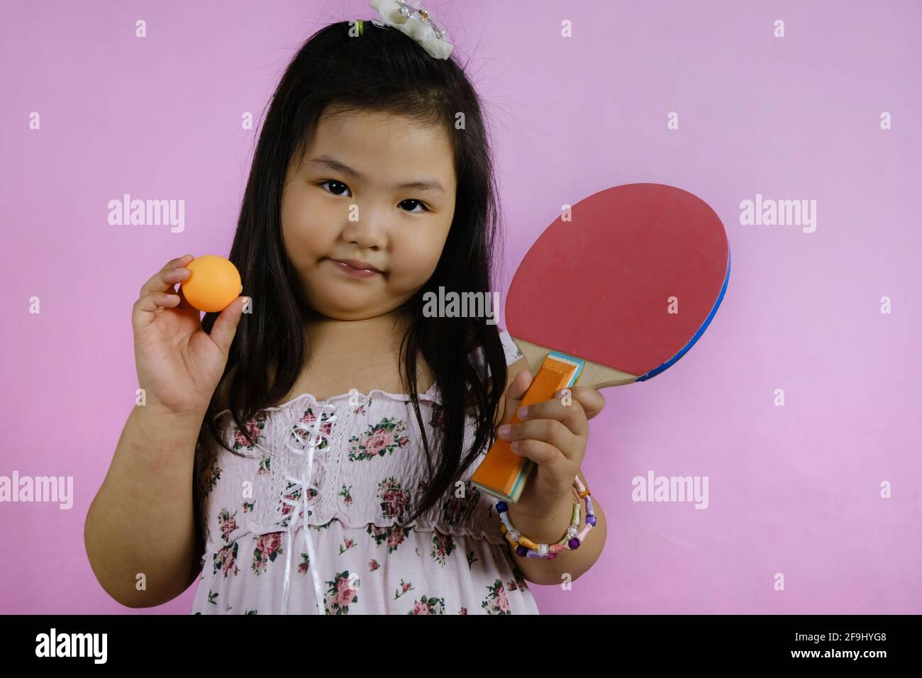 Ein süßes junges molliges asiatisches Mädchen zeigt ihr Tischtennis-Paddel und ihren Ball, macht sich bereit zum Spielen, fühlt sich glücklich und selbstbewusst. Rosa Hintergrund. Stockfoto