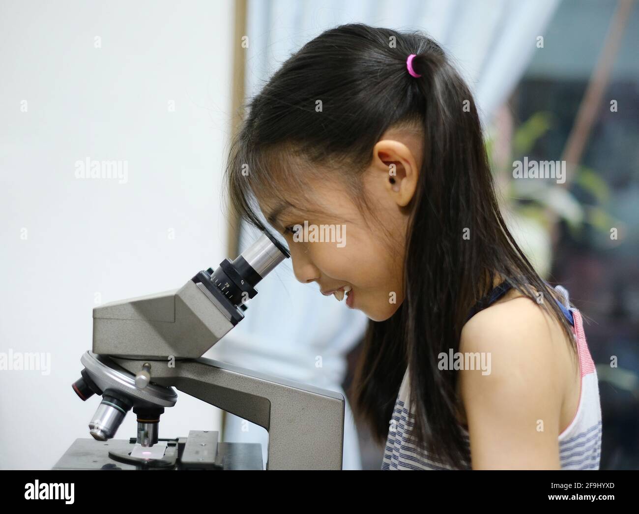 Ein süßes, junges asiatisches Mädchen lernt in ihrem Biologielabor das Mikroskop zu benutzen, macht Spaß und inspiziert einzellige Organismen. Stockfoto