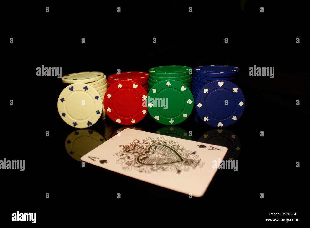 Weiße, rote, grüne, blaue Pocker-Chips stapeln sich mit vier Chips und einer Ass-Karte vorne isoliert auf schwarz mit Reflektion Stockfoto