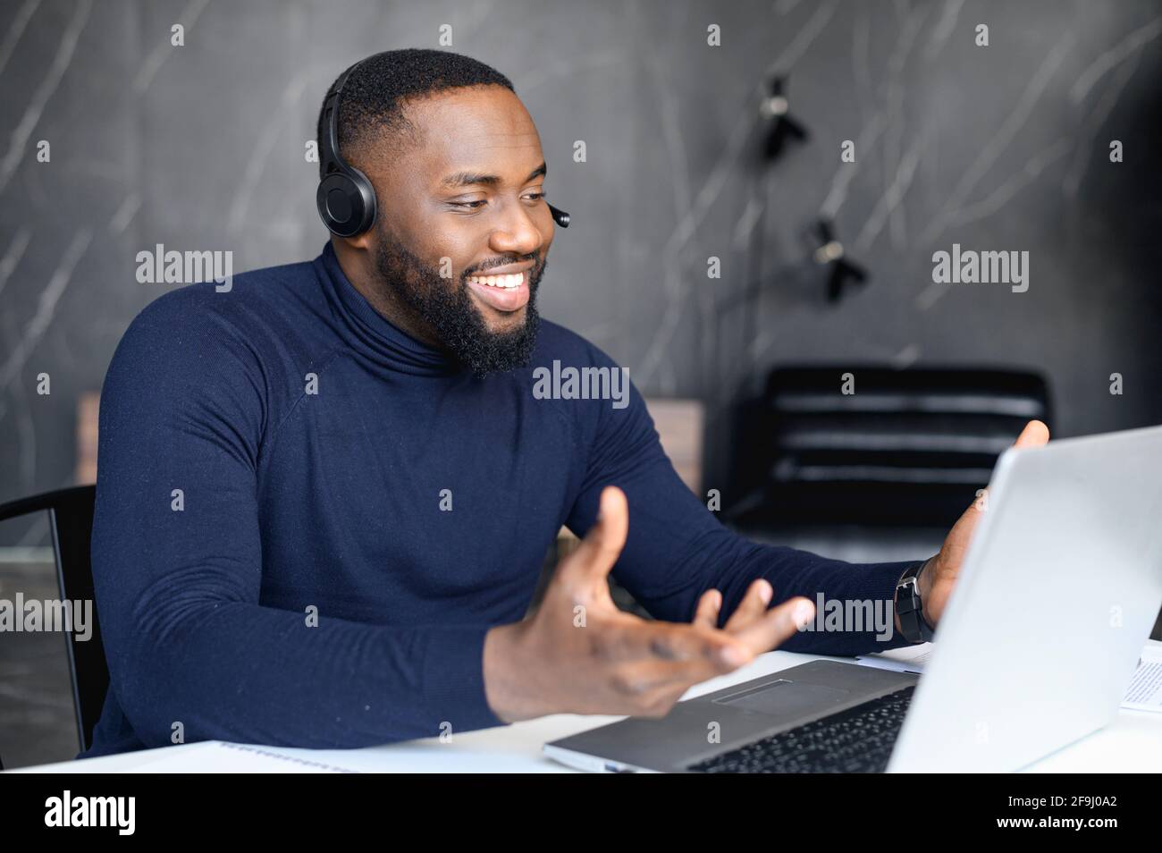 Fokussierter junger Geschäftsmann in einer Brille mit Kopfhörern, der Videoanruf mit Kunden auf dem Laptop hielt. Konzentrierter afrikanischer Mann in einer Brille, der Online-Unterricht hält und Kunden berät Stockfoto