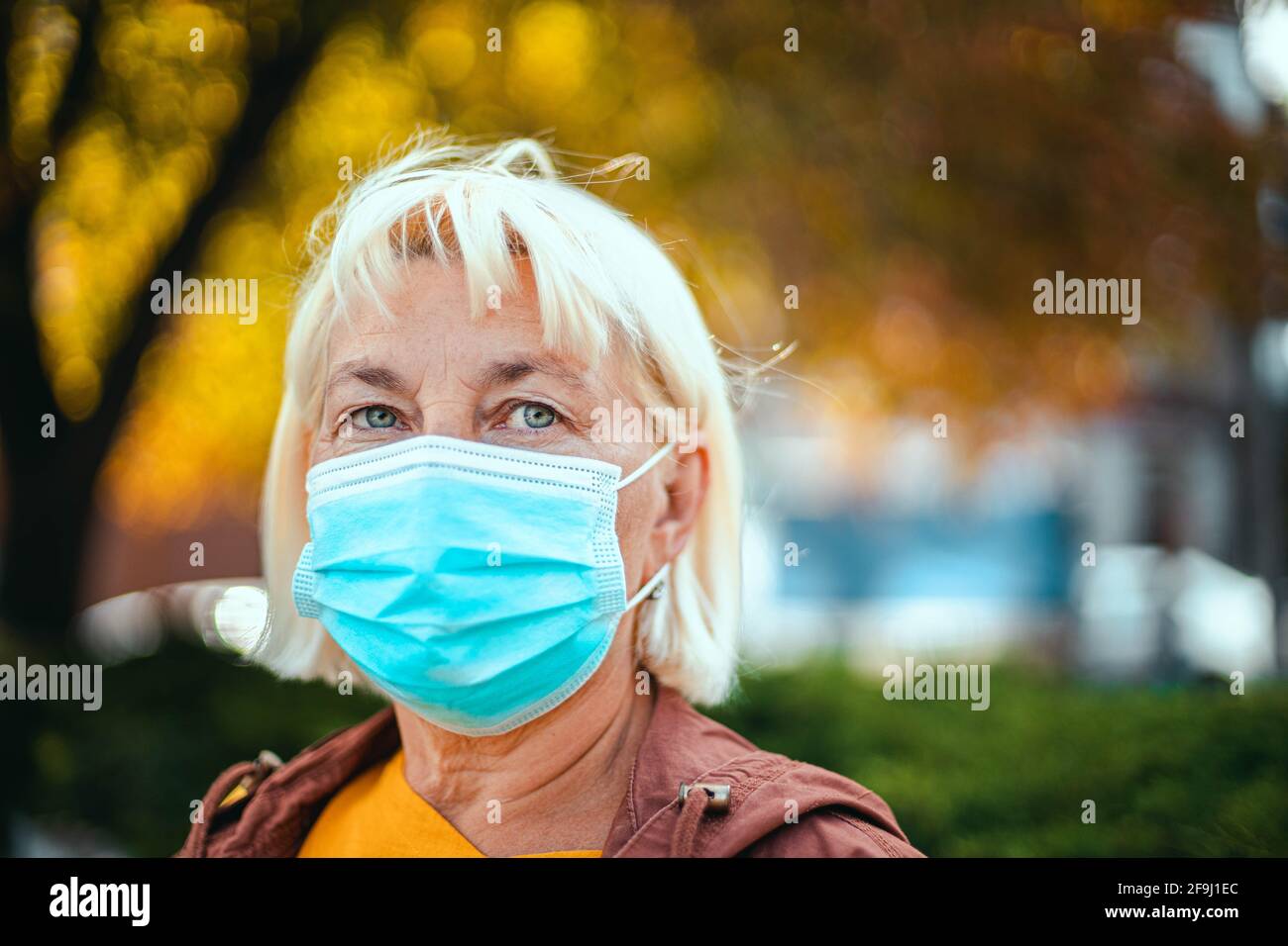 Kaukasische Erwachsene blonde Frau 50 Jahre alt in hellen Gewand Und schützende medizinische Atemschutzmaske weg auf den Park schauen Stockfoto
