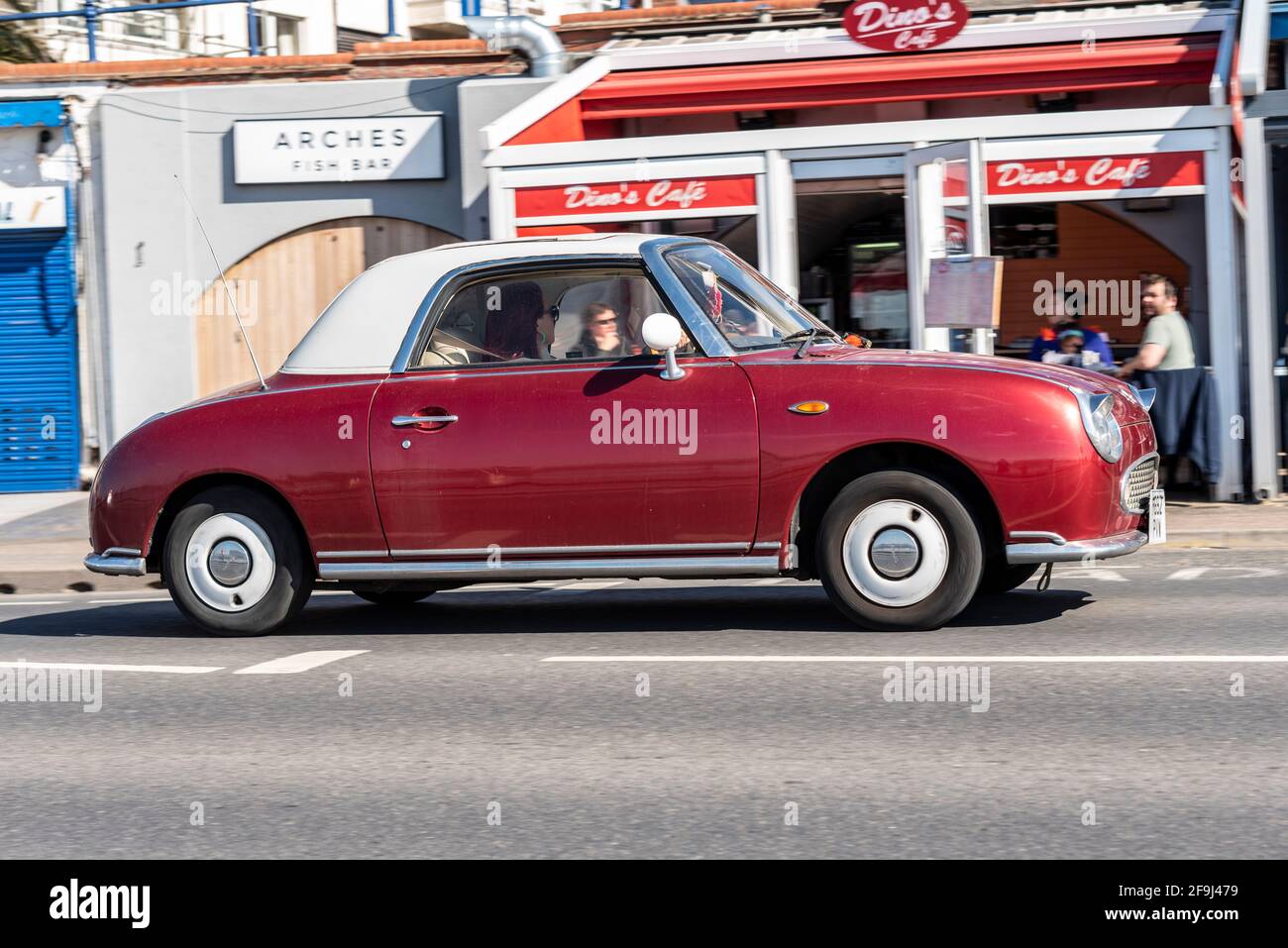 Nissan Figaro fährt in Southend on Sea, Essex, Großbritannien, an einem sonnigen, hellen Frühlingstag, Vorbei an Arches Cafe, Restaurants. Nicht standardmäßige magentafarbene Gehäusefarbe Stockfoto