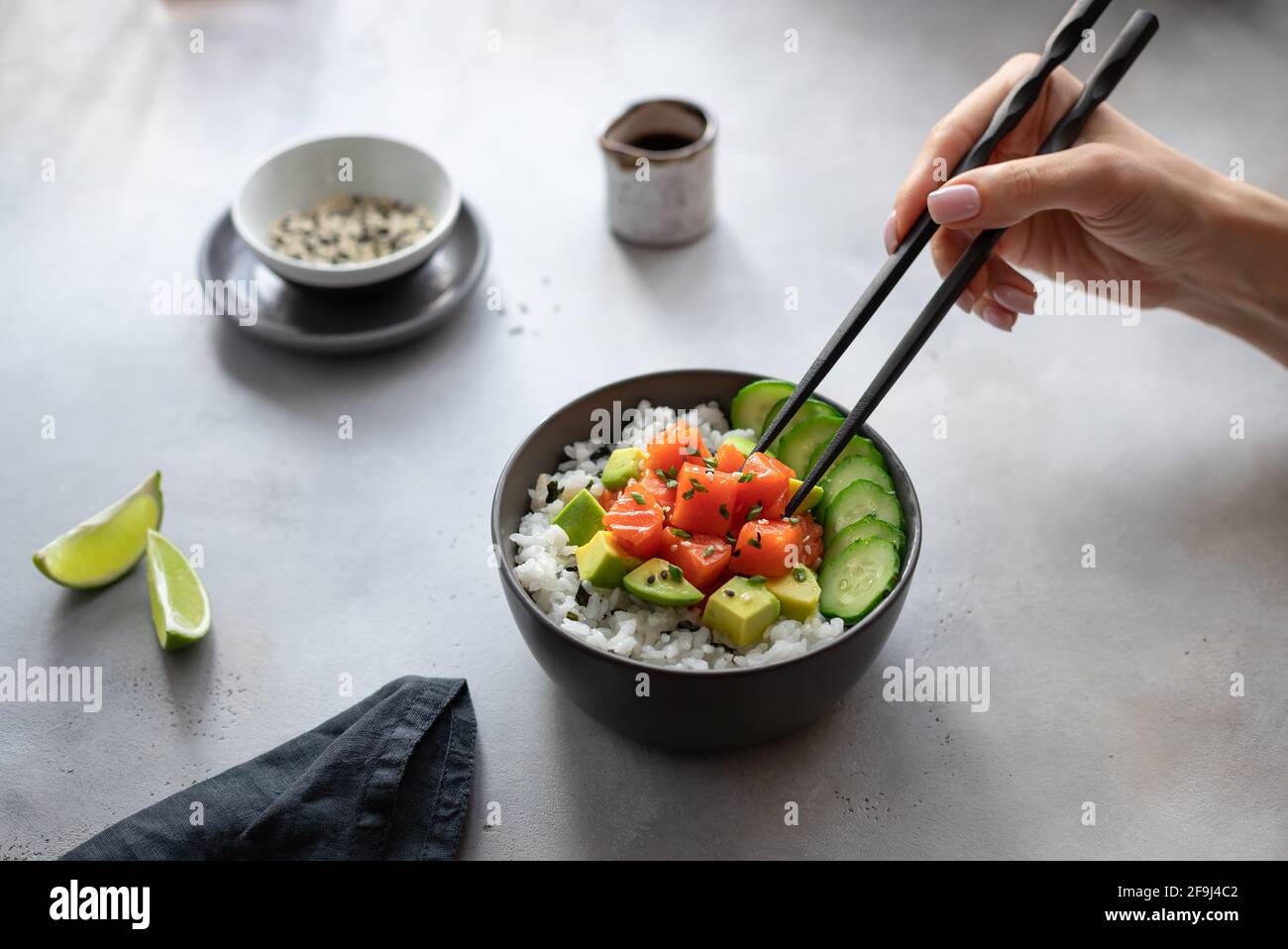 Frau, die Hand isst, Essstäbchen, hawaiianische Schüssel mit Lachs, Reis, Avocado und Gurke. Ernährung. Horizontales Bild, grauer Hintergrund. Stockfoto