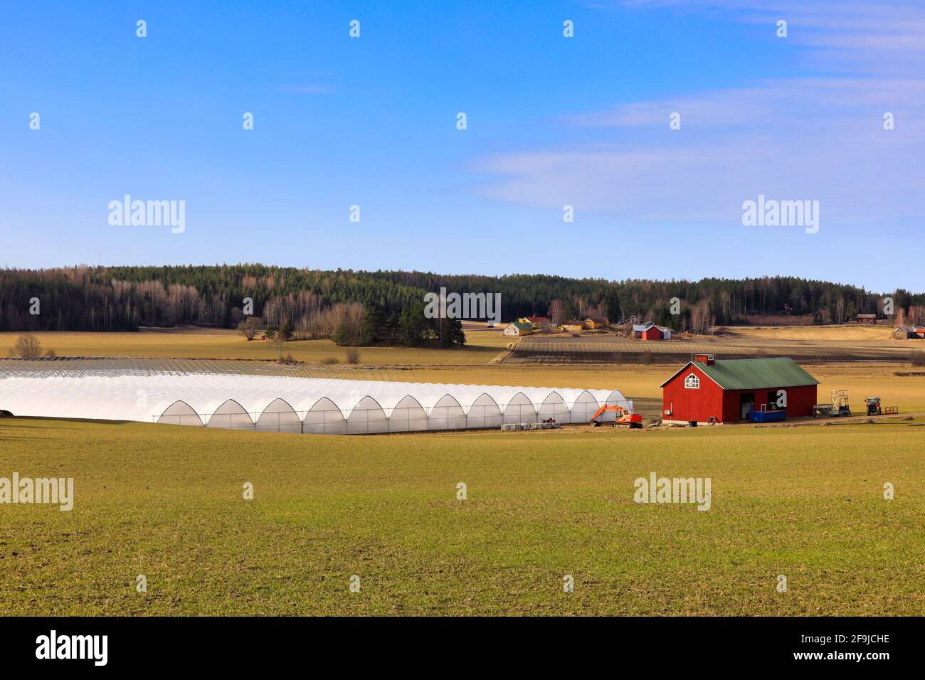 Landwirtschaftliche Landschaft von Gewächshäusern, Feld und roten Stützgebäude mit Maschinen unter blauem Himmel mit einigen Wolken. Sauvo, Finnland. 4. April 2021 Stockfoto