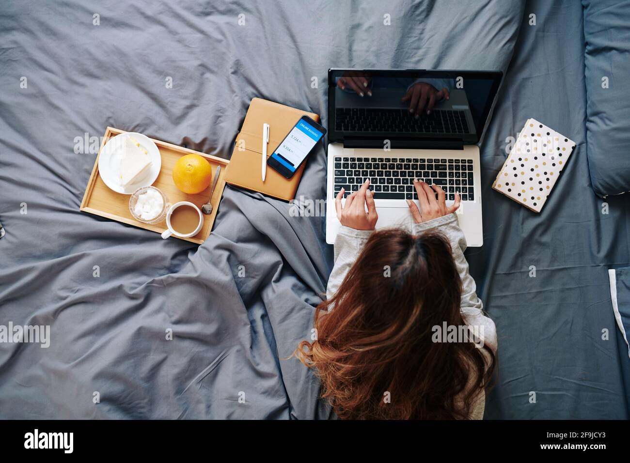 Hübsche junge Frau, die im Bett am Laptop arbeitet, Frühstück isst und ihr Bankkonto über die Anwendung auf dem Smartphone überprüft Stockfoto