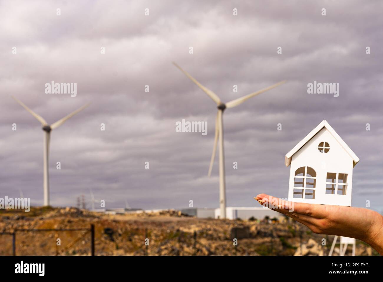 Hand einer Frau, die ein weißes Holzhausmodell in der Nähe von Windenergieanlagen hält. Konzept für grüne, nachhaltige und erneuerbare Energien. Stockfoto