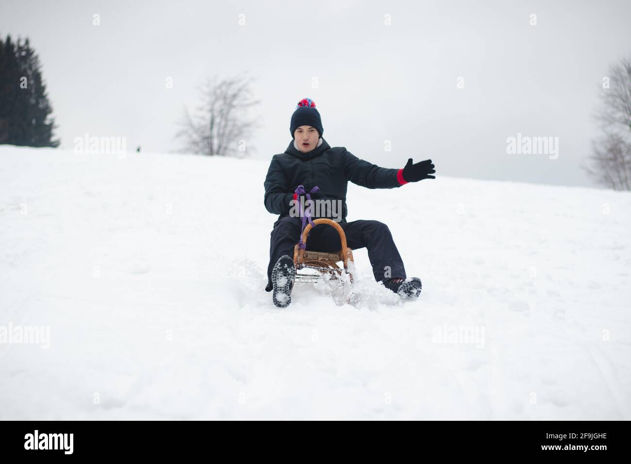 Erwachsener Mann in winterlicher schwarzer Kleidung genießt mit einem Lächeln eine Fahrt auf einem Holzschlitten und versucht nicht davon zu fallen. Fußbremsung. Verwendung von frischem Pulver für ein Stockfoto
