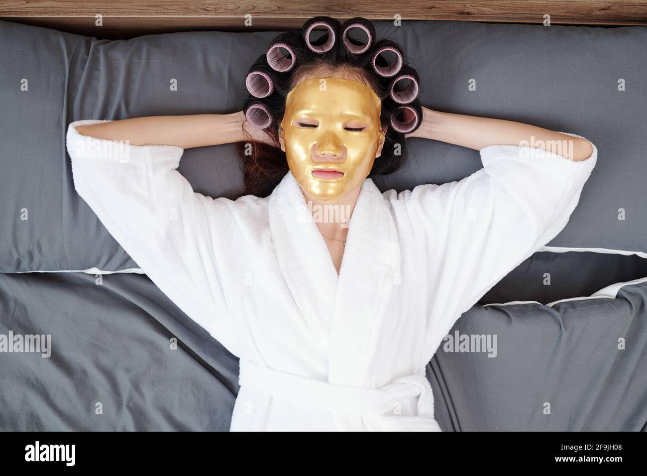 Junge Frau im Bademantel entspannt auf dem Bett mit Haarwalzen und Bettmaske auf, Blick von oben Stockfoto