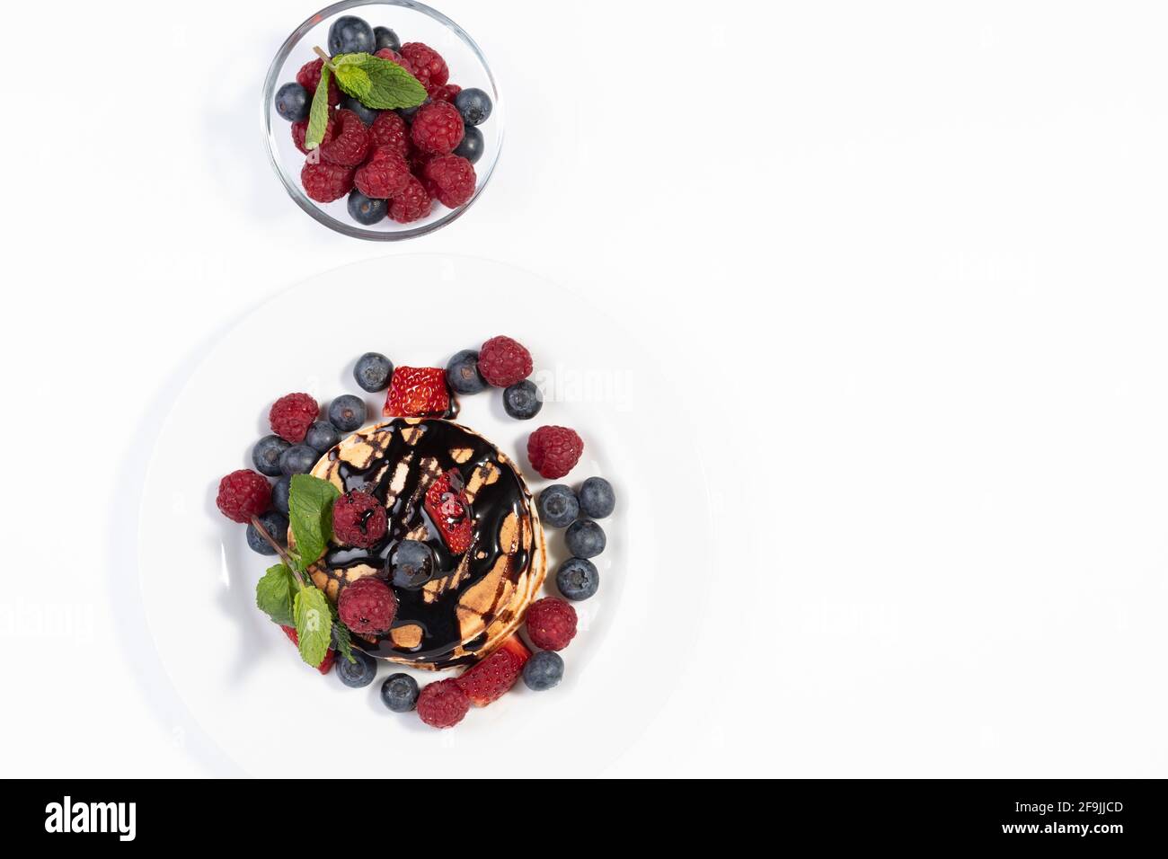 Draufsicht auf Pfannkuchen mit Schokoladensirup, Erdbeeren, Himbeeren, Heidelbeeren und Minzblättern auf weißem Teller auf weißem Hintergrund. Teller mit Scheibe Stockfoto