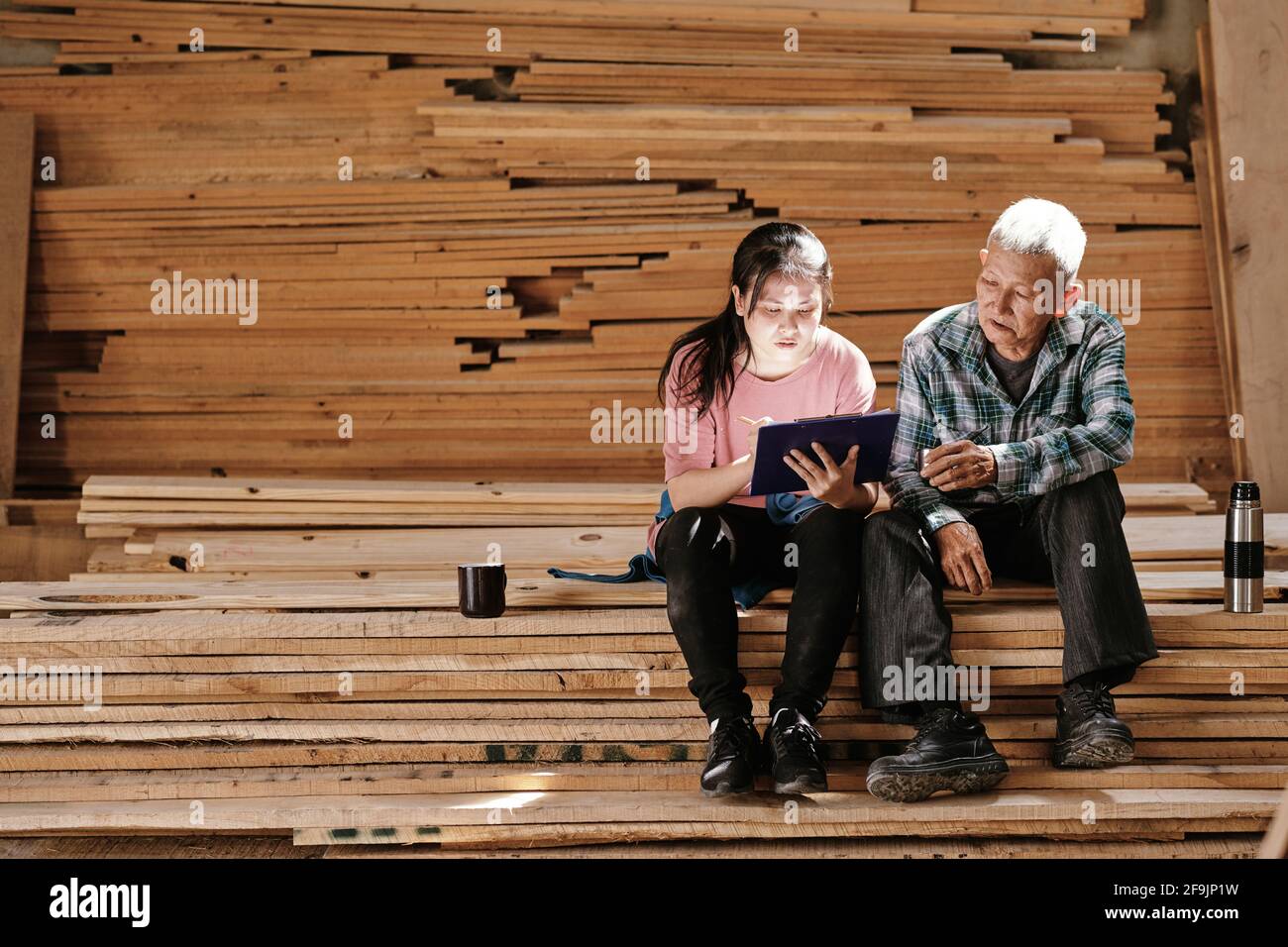 Großvater und Enkelin ruhen sich auf gestapelten Holzbrettern aus und arbeiten Auf Skizze des Projekts für den Kunden Stockfoto