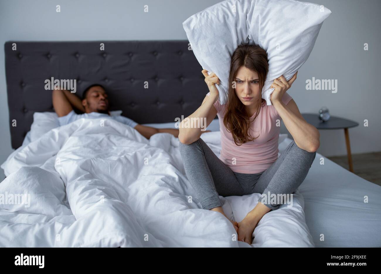 Verärgert junge Frau, die Ohren mit Kissen bedeckt, um das Schnarchen ihres Mannes auf dem Bett nicht zu hören, leerer Raum Stockfoto