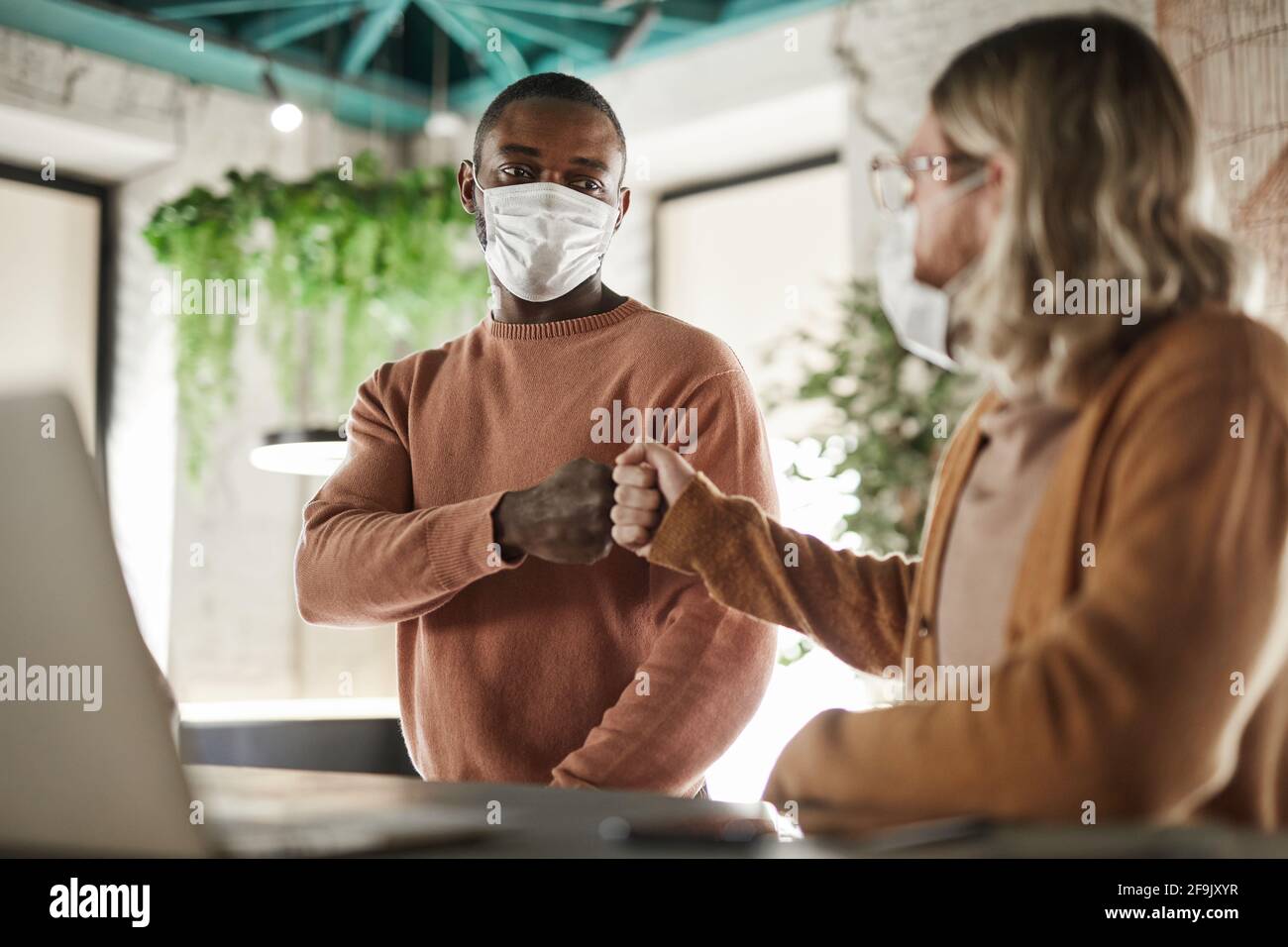 Portrait von zwei Männern, die Masken und anstoßende Fäuste tragen, während einer taktlosen Begrüßung im Café oder Büro, covid Konzept Stockfoto