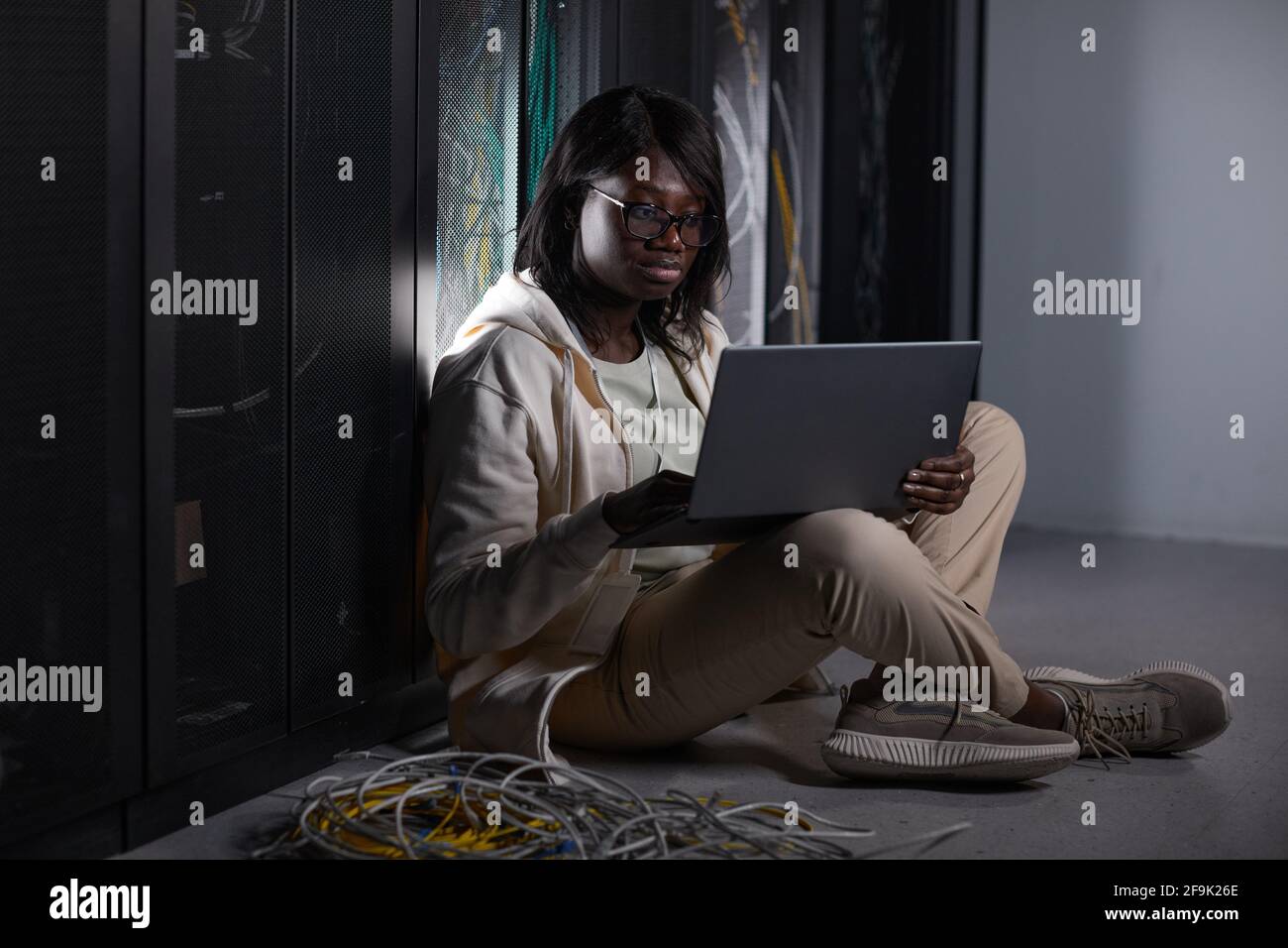 Porträt einer jungen afroamerikanischen Frau, die im Serverraum einen Laptop benutzt und auf dem Boden sitzt, während sie mit einem Supercomputer im Rechenzentrum arbeitet, Stockfoto