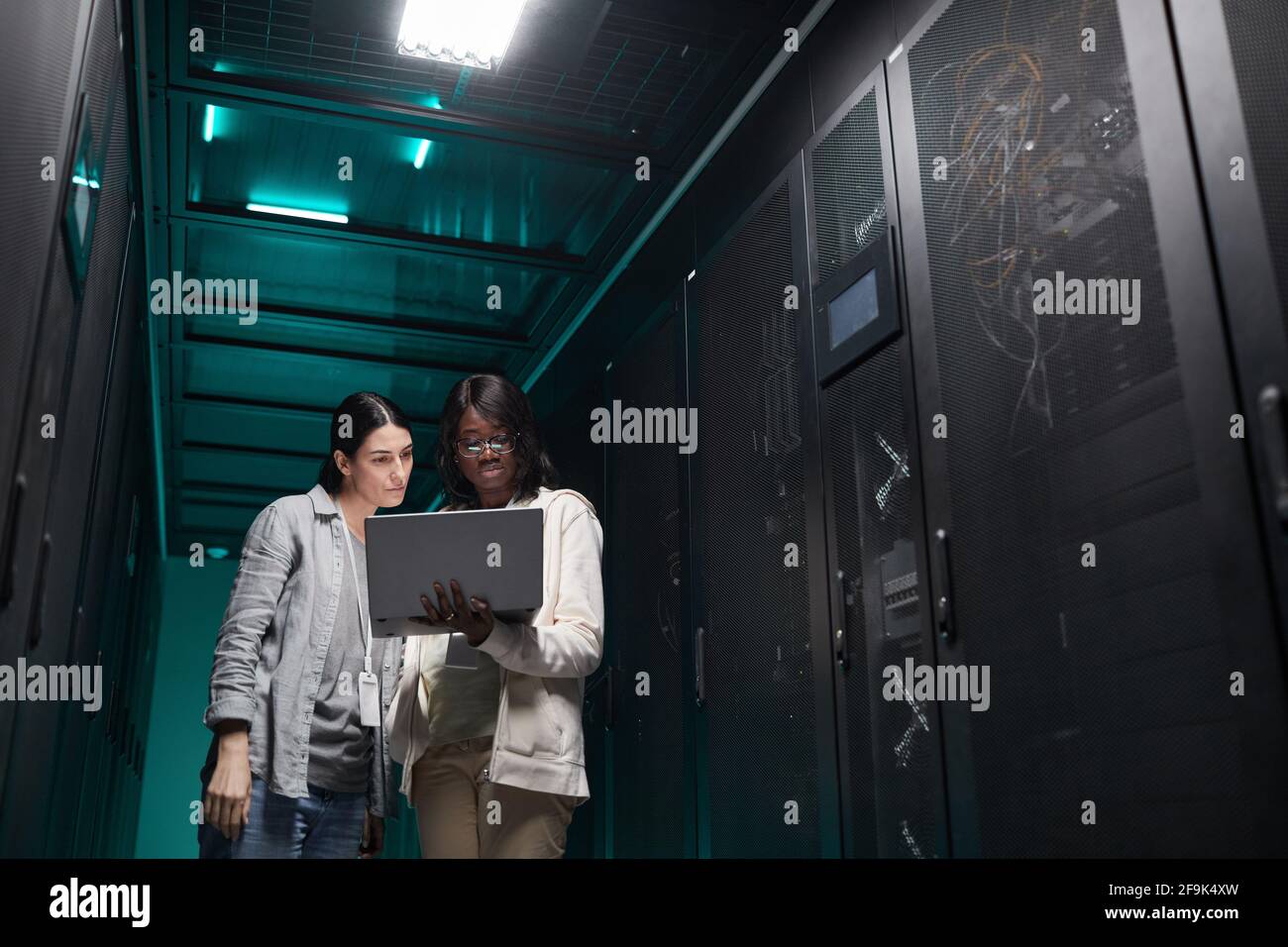 Porträt von zwei weiblichen IT-Technikerinnen, die das Servernetzwerk über einen Laptop einrichten, während sie im Rechenzentrum arbeiten und Platz kopieren Stockfoto