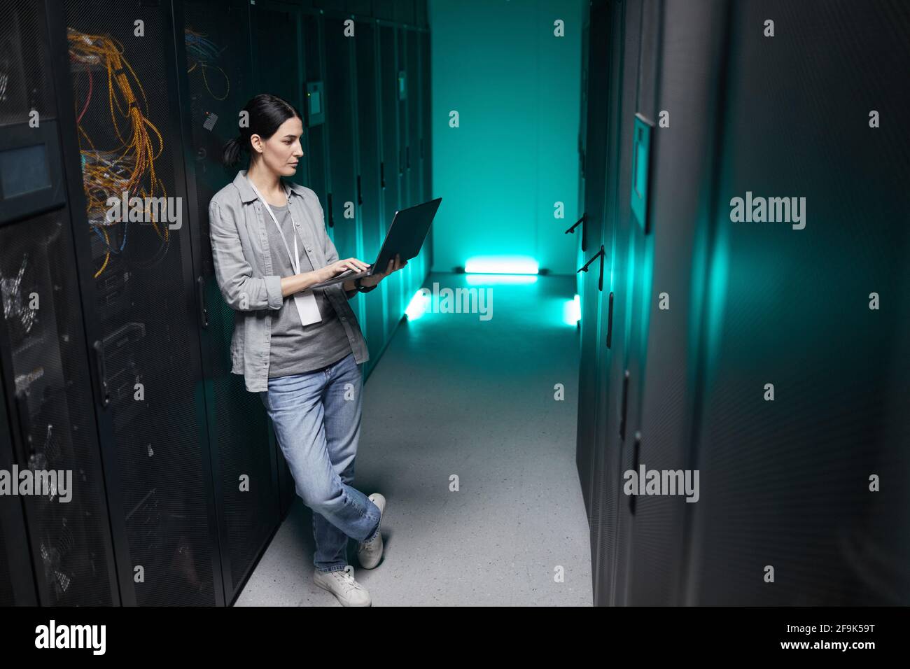 In voller Länge ein Hochformat mit einer Frau, die einen Laptop in der Hand hält, während sie mit einem Supercomputer im Serverraum arbeitet und Platz für Kopien hat Stockfoto