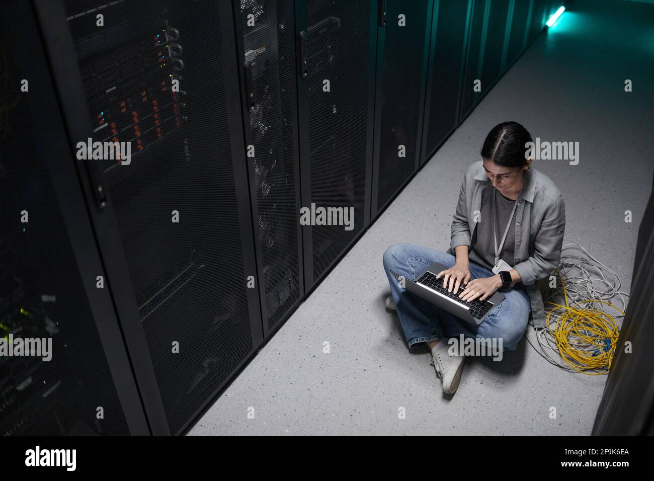 Eine Frau mit einem Laptop, die im Serverraum auf dem Boden sitzt und ein Supercomputer-Netzwerk aufstellt, kann aus einem hohen Blickwinkel betrachtet werden Stockfoto