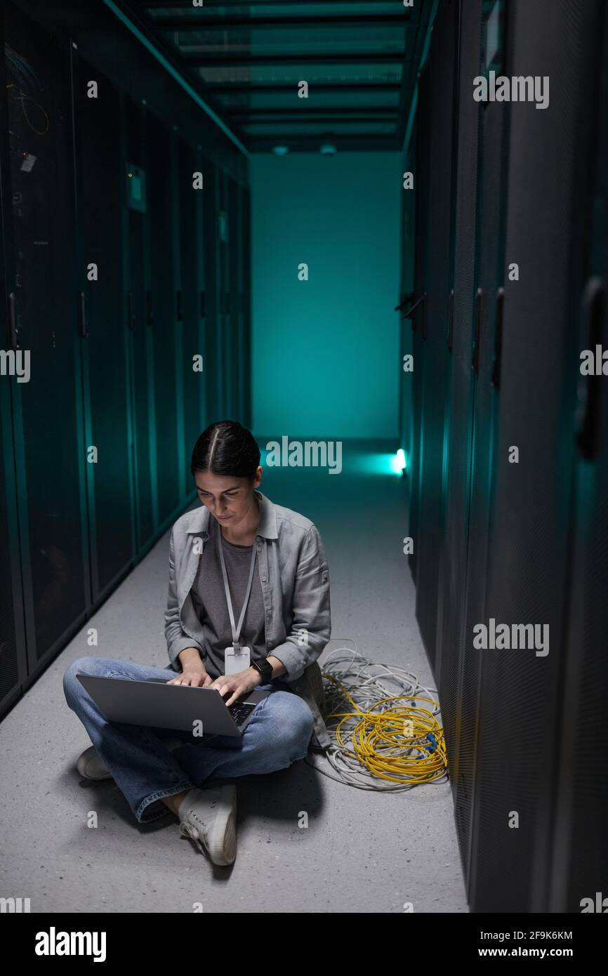 Vertikales Porträt einer Frau, die einen Laptop benutzt, während sie im Serverraum auf dem Boden sitzt und ein Supercomputer-Netzwerk aufstellt und Platz kopiert Stockfoto