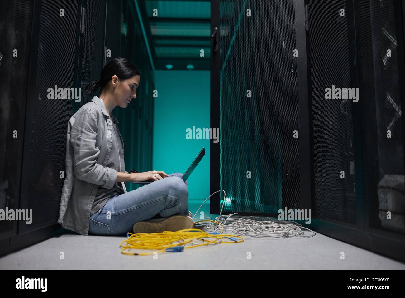 Seitenansicht Porträt einer Frau, die einen Laptop benutzt, während sie im Serverraum auf dem Boden sitzt und ein Supercomputer-Netzwerk aufstellt und Platz kopiert Stockfoto