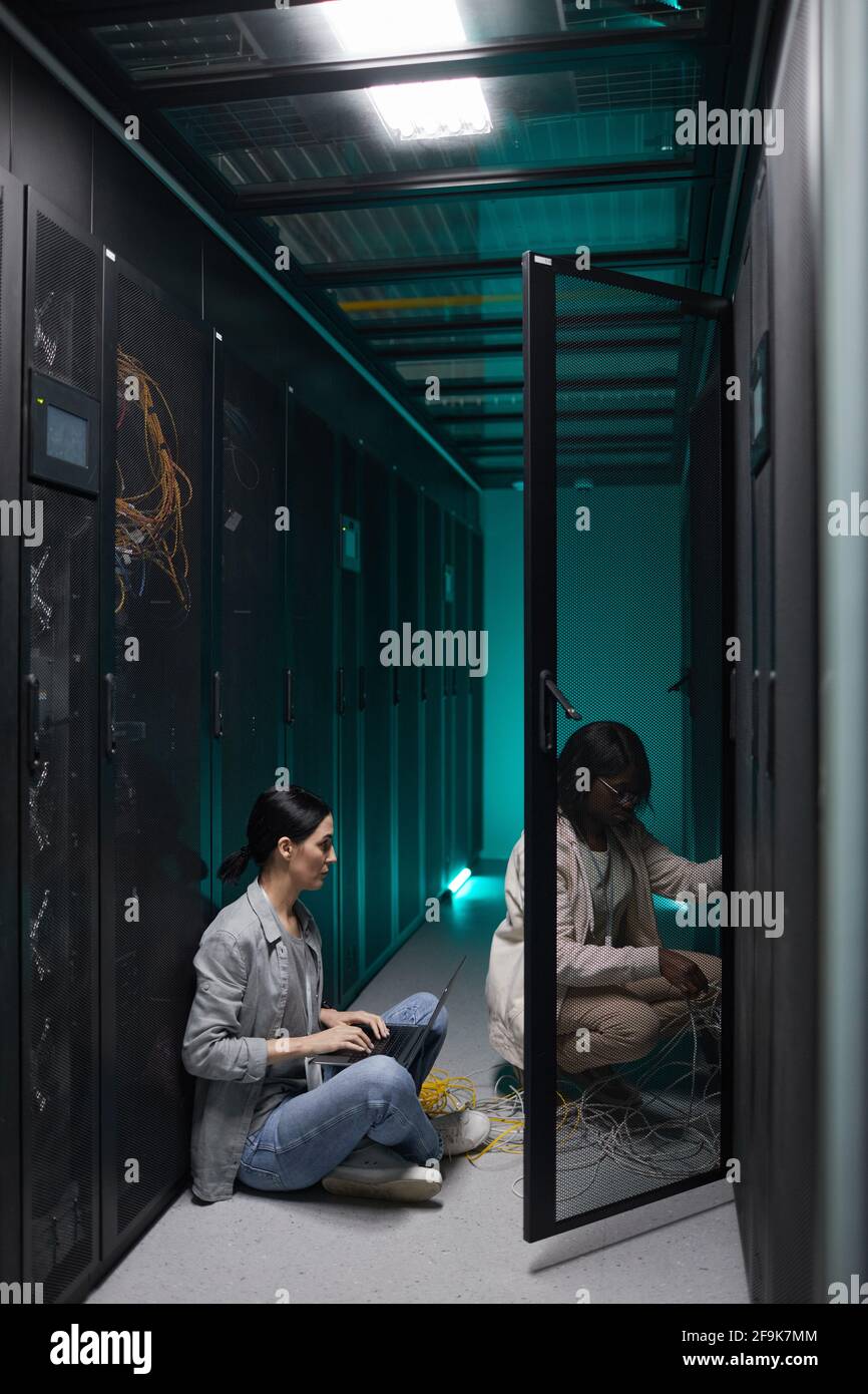 Vertikales Porträt zweier weiblicher Dateningenieure, die einen Laptop im Serverraum verwenden und ein Supercomputer-Netzwerk einrichten und Speicherplatz kopieren Stockfoto