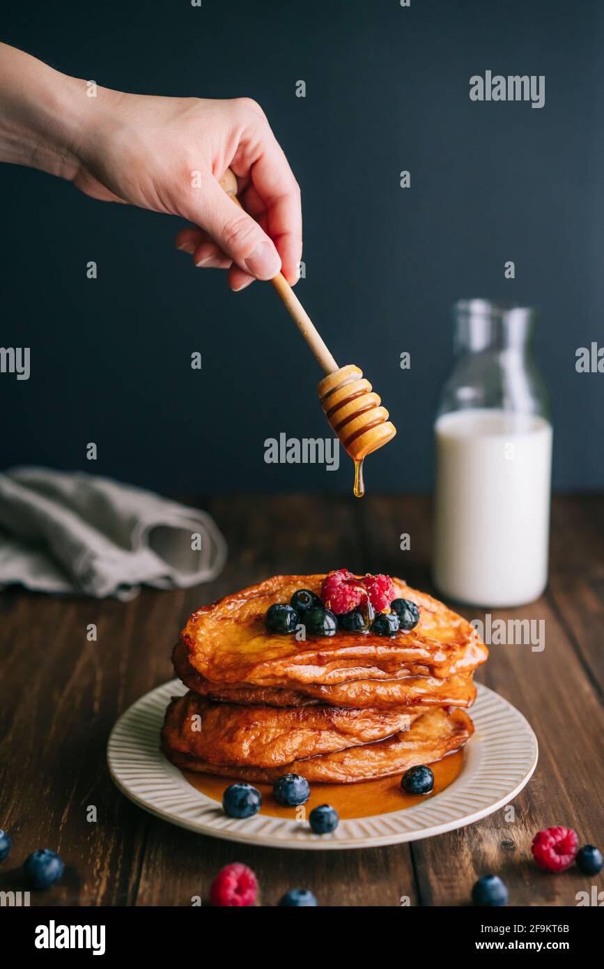 Hand mit Honiglöffel gießt Honig über gebratene Brotscheiben mit Blaubeeren und Himbeeren auf einem alten beigen Teller auf einen Holztisch. Spanische Torrijas Stockfoto