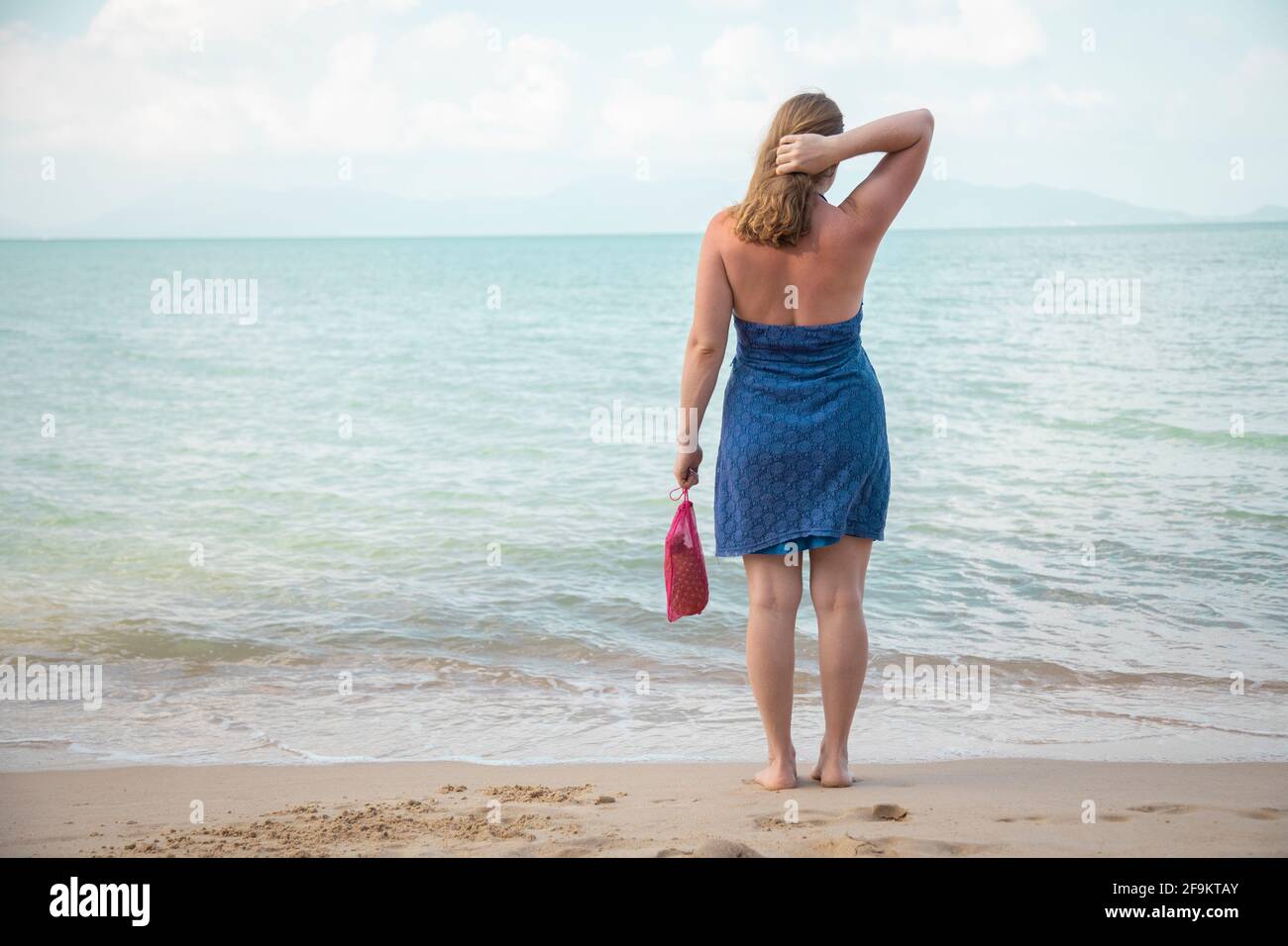 Eine Frau hält in der Hand eine Mesh-Einkaufstasche und steht am Rande des Meeres an einem Sandstrand. Ökologie-Konzept. Bewusster Konsum Stockfoto