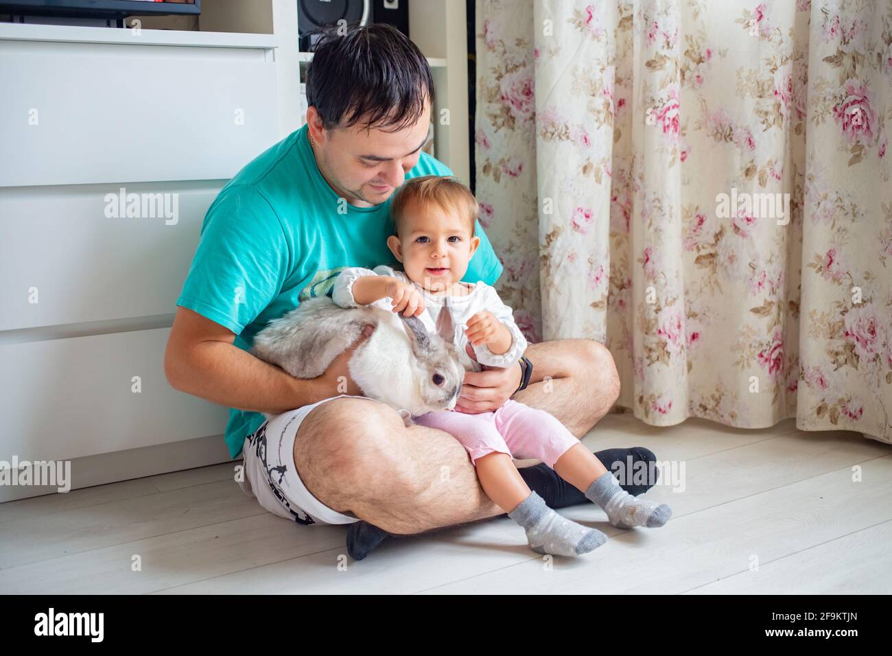 Entzückendes Baby sitzt in den Armen des Vaters und streichelt ein dekoratives Kaninchen. Vater zeigt kleines Kind osterhase. Haustiere in einer Familie mit Kindern Stockfoto