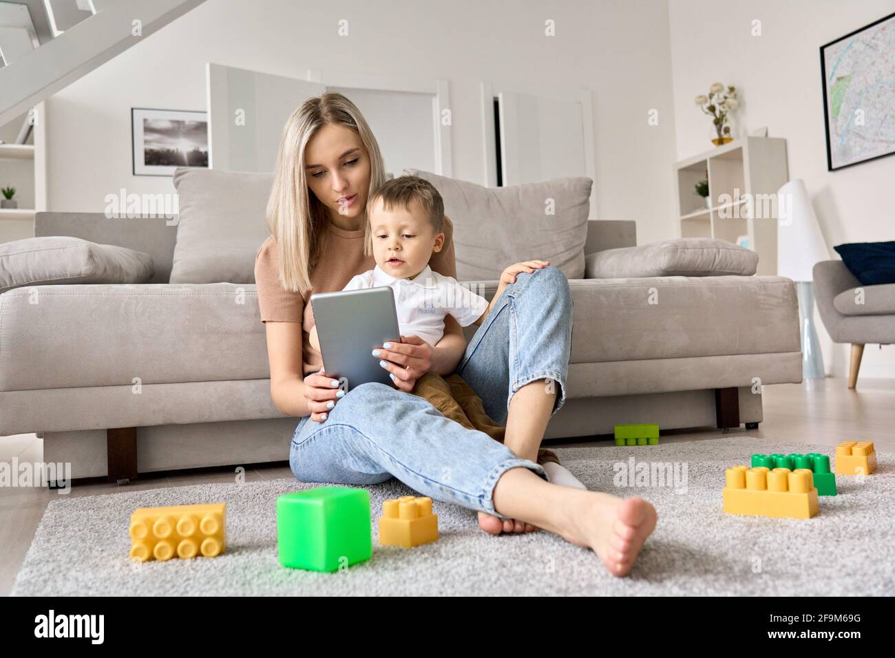 Junge Mutter, die zu Hause auf dem Boden sitzt, während das Kind Filme auf dem Tablet ansieht. Stockfoto