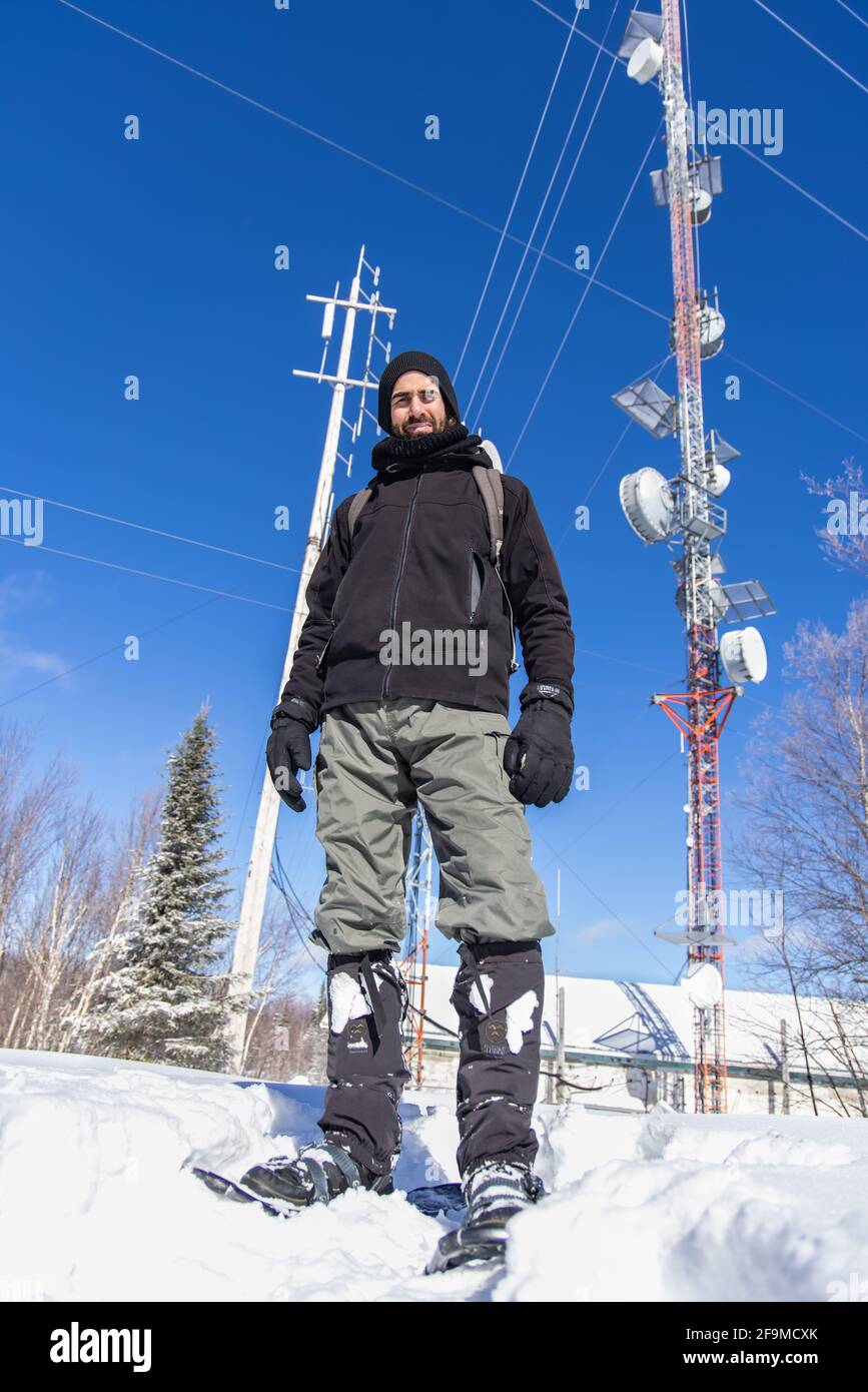 Kippbare Aufnahme eines männlichen Touristen, der vor Mobilfunkmasten mit verschiedenen Antennen posiert, die für die 3G-, 4G- und 5G-Netzkommunikation verwendet werden. Stockfoto