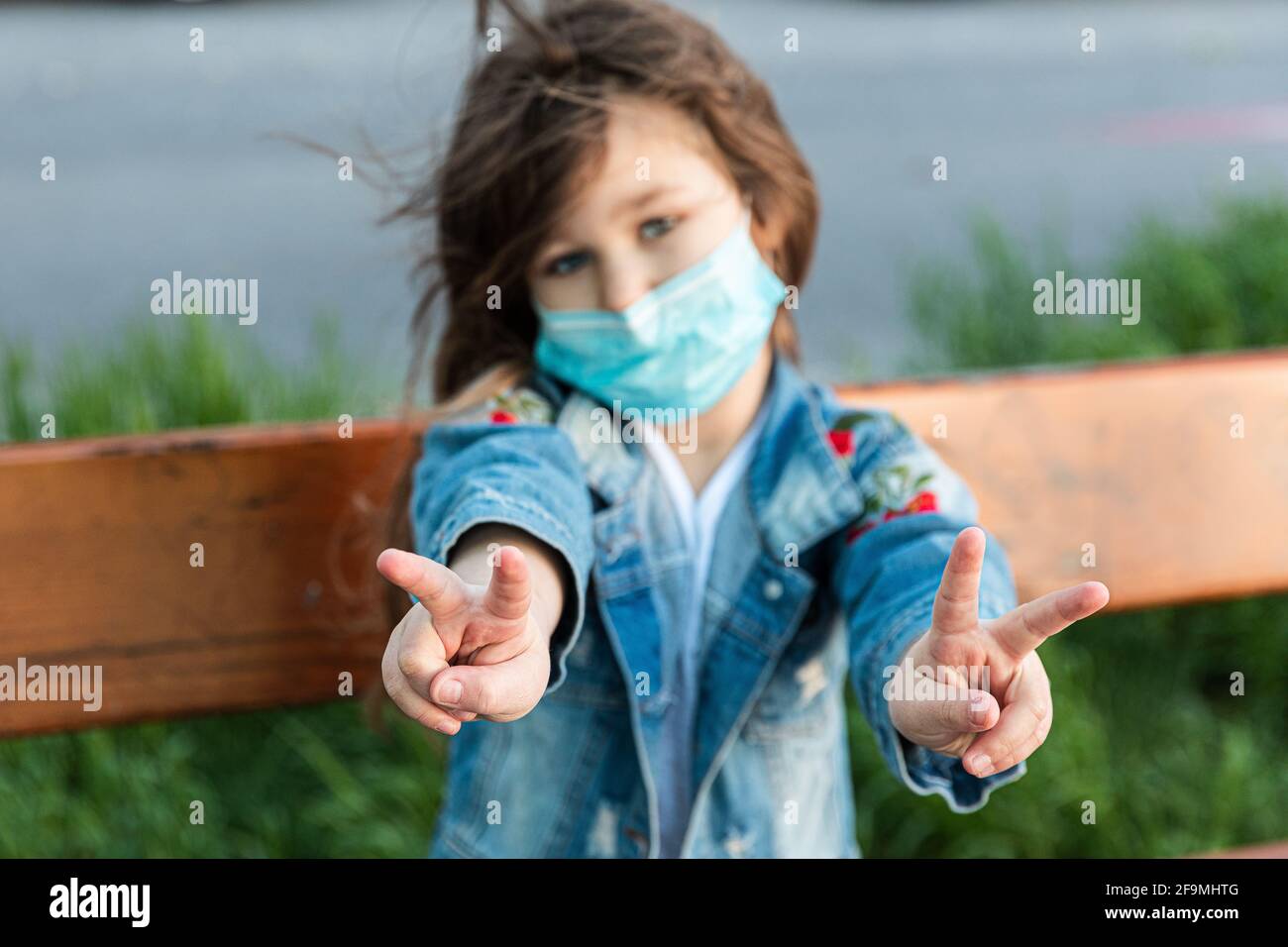 Kaukasisches Mädchen, das auf einer Bank in der Maske sitzt Das Gesicht mit traurigen Augen und zeigt Hände Geste Viva Stockfoto