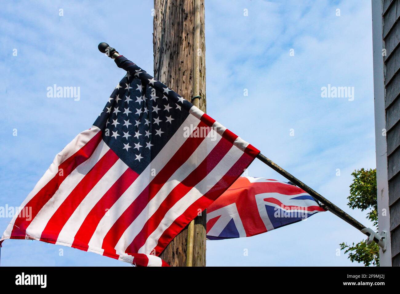 Britische und amerikanische Flaggen fliegen von einem verwitterten Klappbretthaus Vor einem elektrischen Pol und einem blau bewölkten Himmel Stockfoto