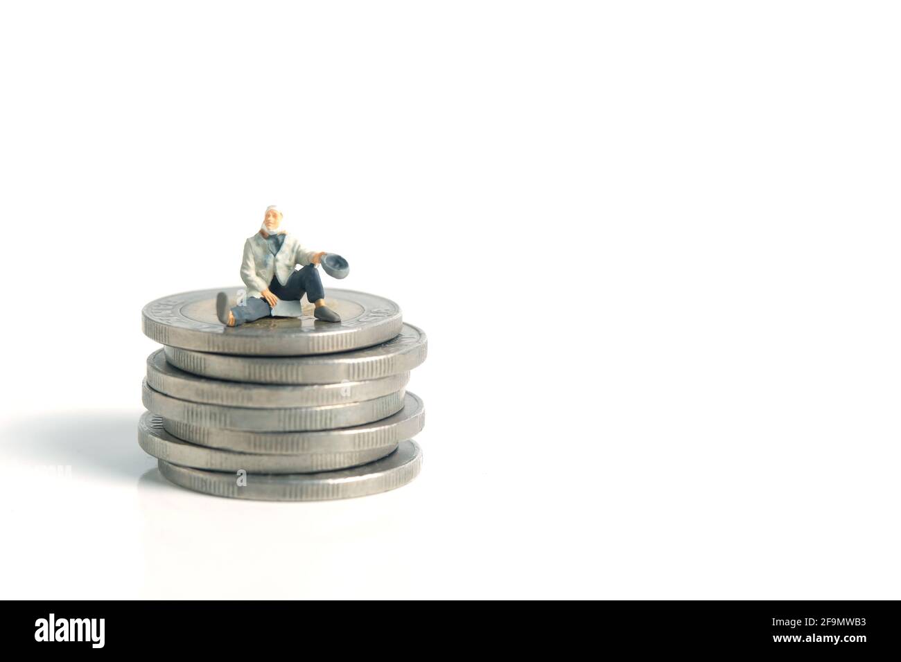Miniatur kleine Menschen Spielzeug Fotografie. Ein armer Mann oder ein Bettler sitzen über dem Geldmünzstapel, isoliert auf weißem Hintergrund Stockfoto