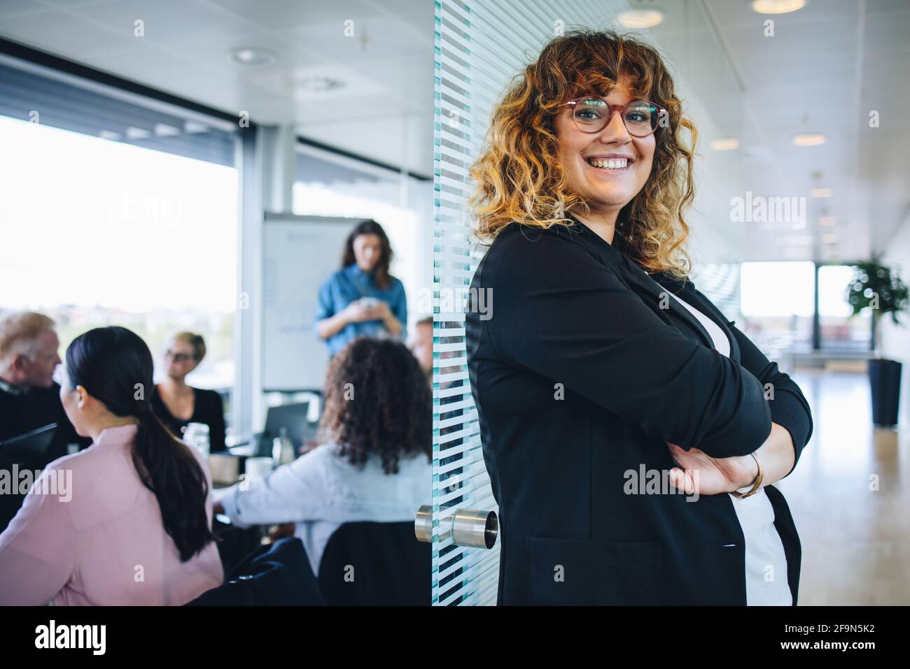 Porträt einer lächelnden Geschäftsfrau, die im Büro mit Kollegen im Besprechungsraum steht. Erfolgreiche weibliche Profikerin mit gekreuzten Armen. Stockfoto