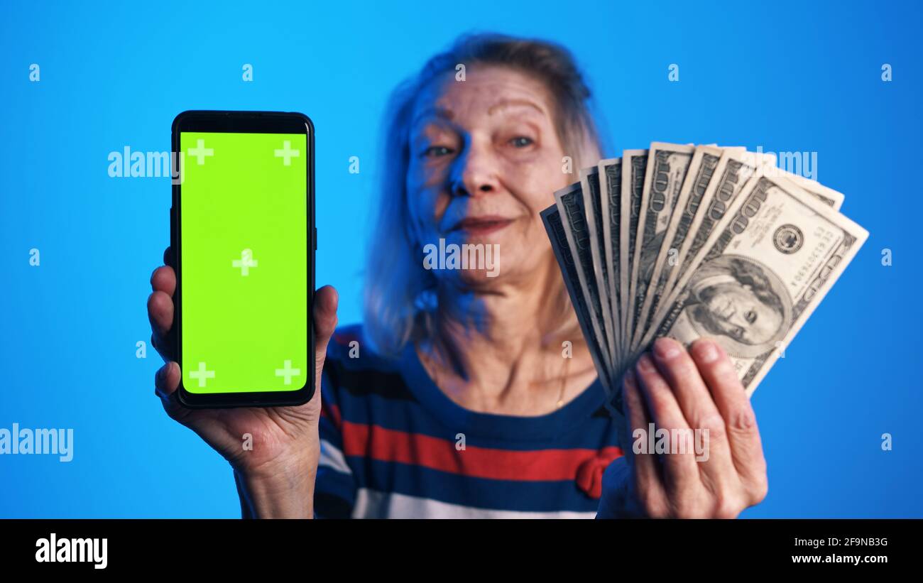 Ältere Frau hält Telefon mit grünem Bildschirm in der einen und Geld in der anderen Hand. Lotteriegewinn. Hochwertige Fotos Stockfoto