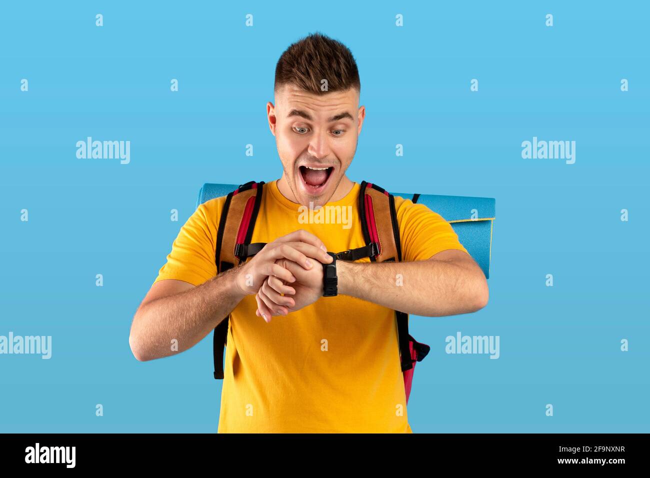 Besorgter junger Mann mit Campingausrüstung, der seine Uhr ansah und die Zeit über dem blauen Studiohintergrund überprüfte Stockfoto