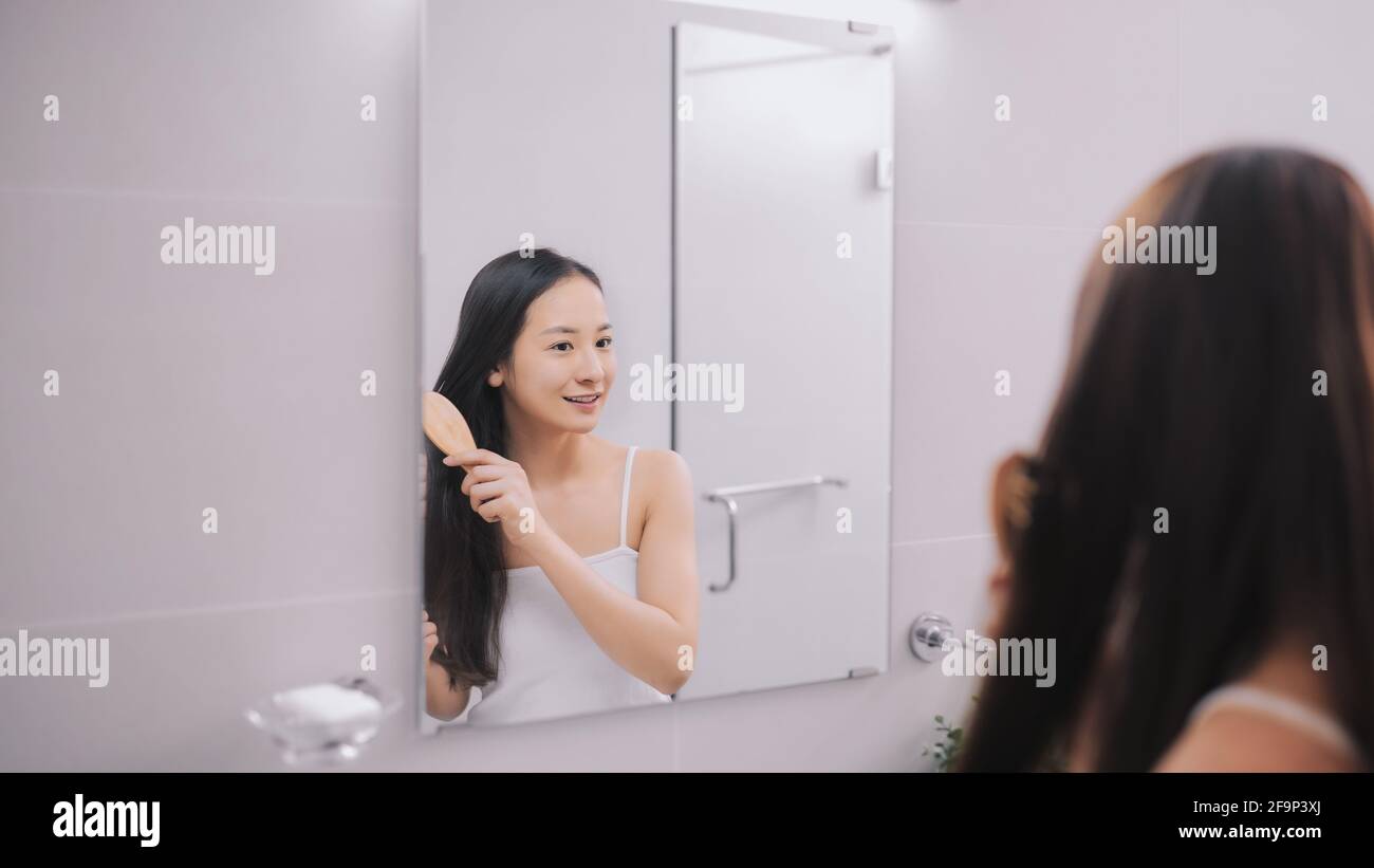 Schöne junge Frau in weißem Unterhemd kämmt ihre Haare und lächelt, während sie in den Spiegel im Badezimmer schaut Stockfoto