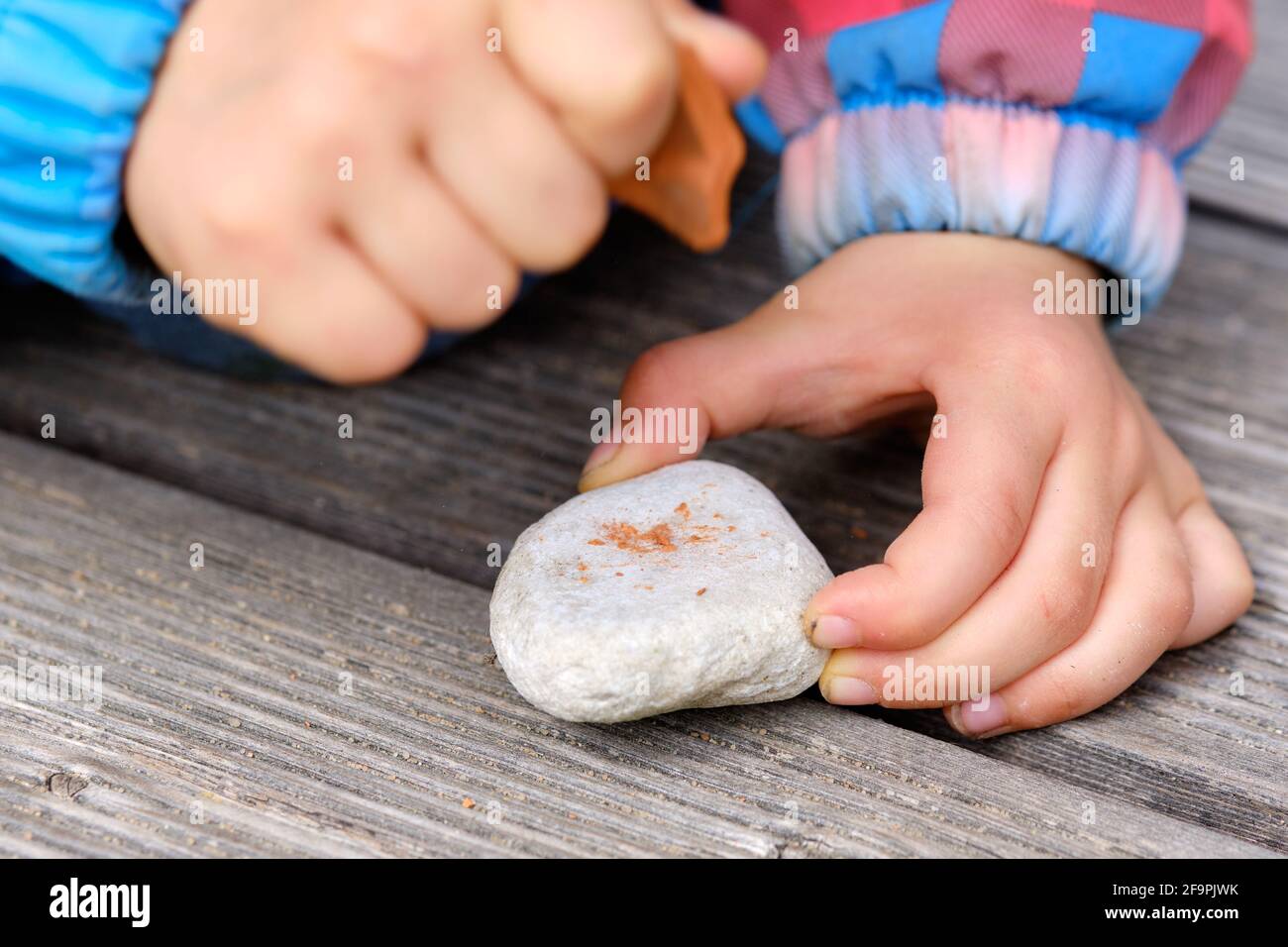 Nahaufnahme der Hände eines 4-jährigen Kindes, das Steine in den Händen hält und sie auf einem Holzboden im Freien aufeinander trifft. Gesehen in Deutschland in Stockfoto