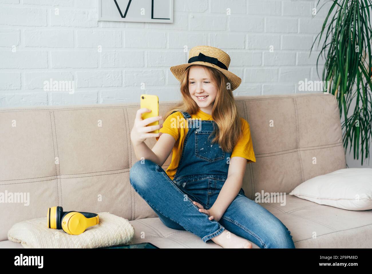 Fröhliches Rotschopf-Mädchen in einem Strohhut und Denim-Overalls, die posieren und ein Selfie auf einem Smartphone machen. Stockfoto